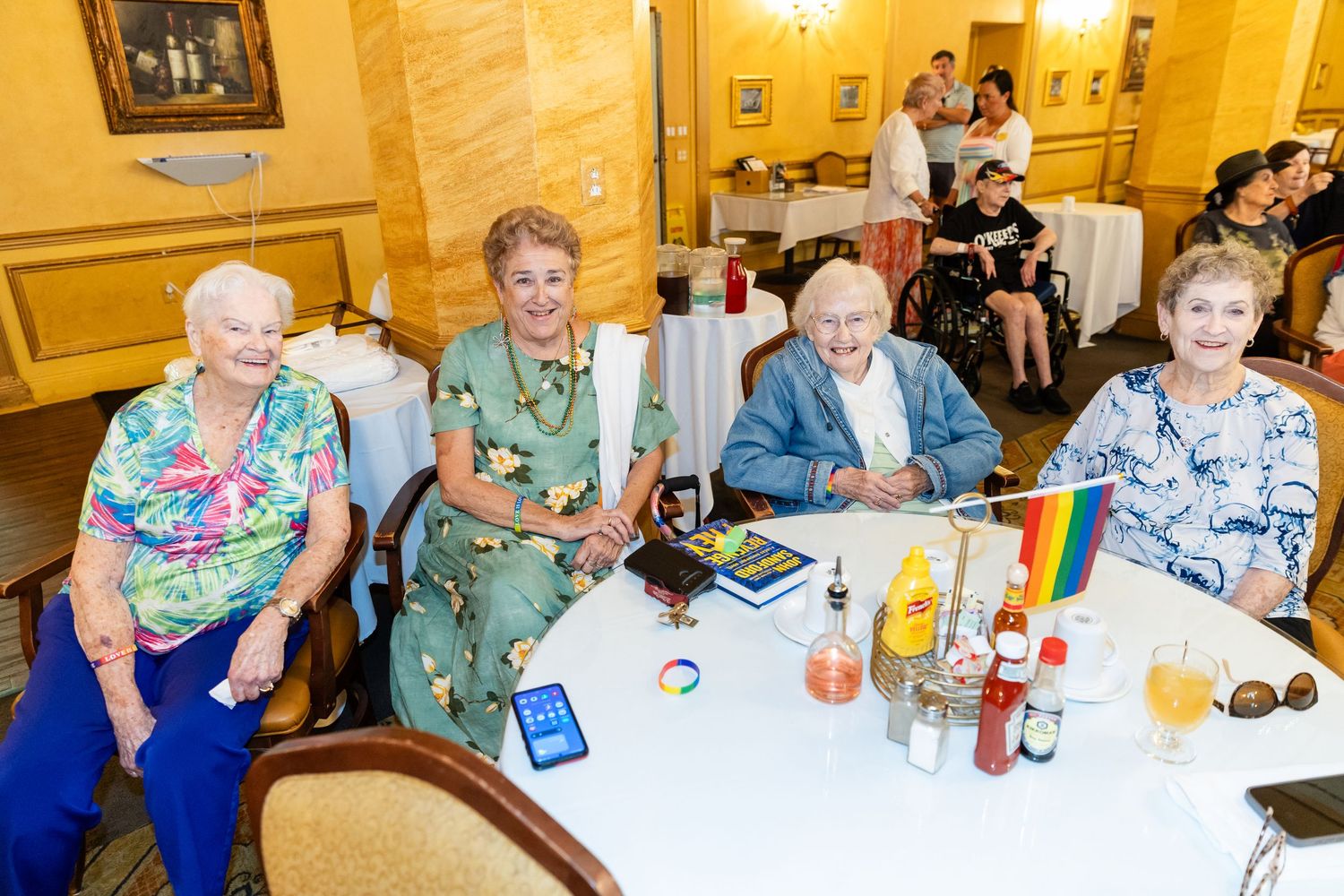 Four elderly adults smile at a decorated dining table with rainbow pride flag during an LGBTQ senior community celebration.