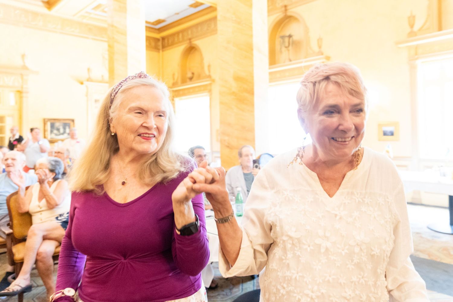 Two smiling older women in purple and white dresses making hand gestures at an indoor celebration.