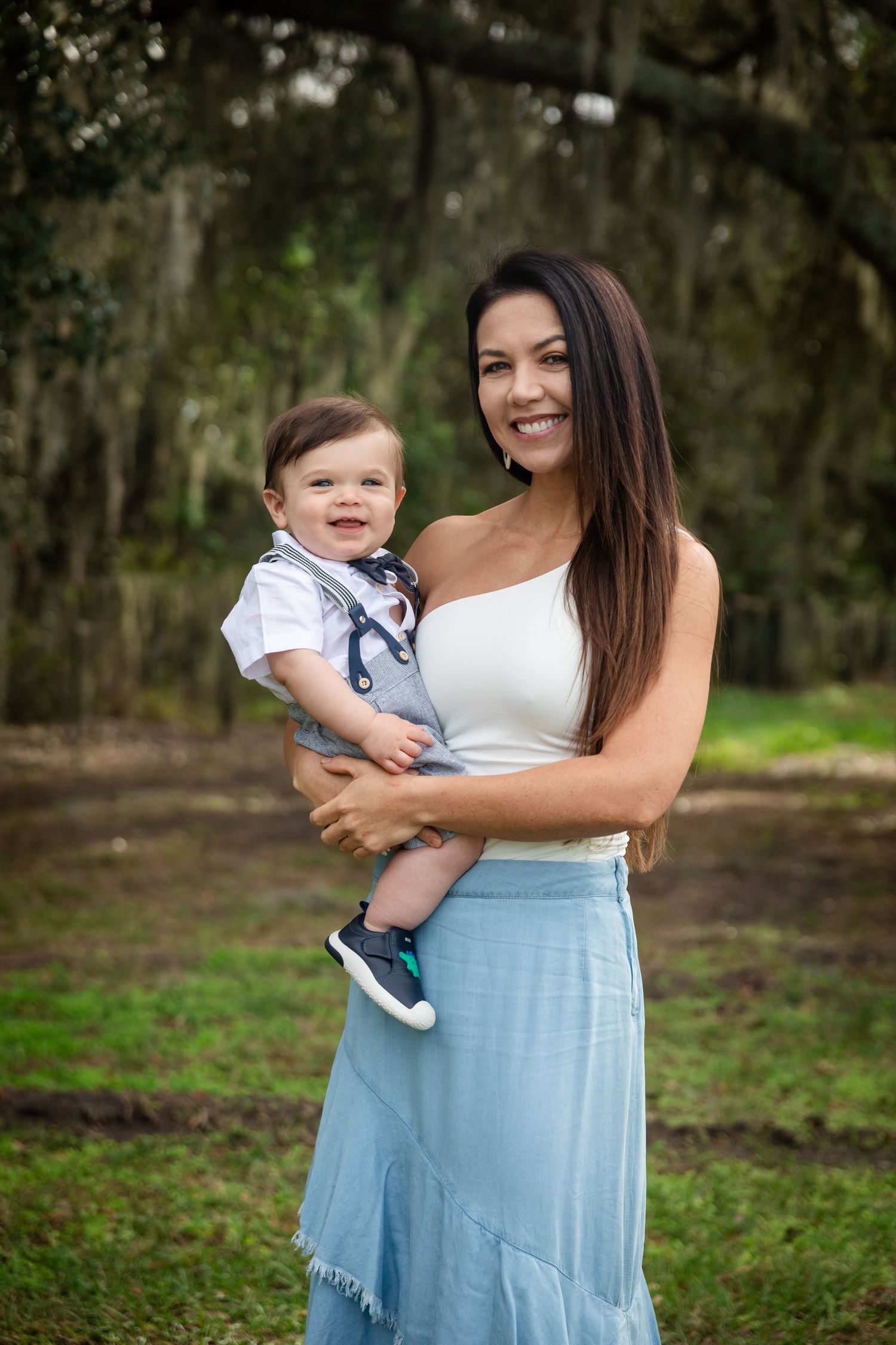 A smiling woman in a white top and blue skirt holds a happy baby boy outdoors in a lush green park setting.