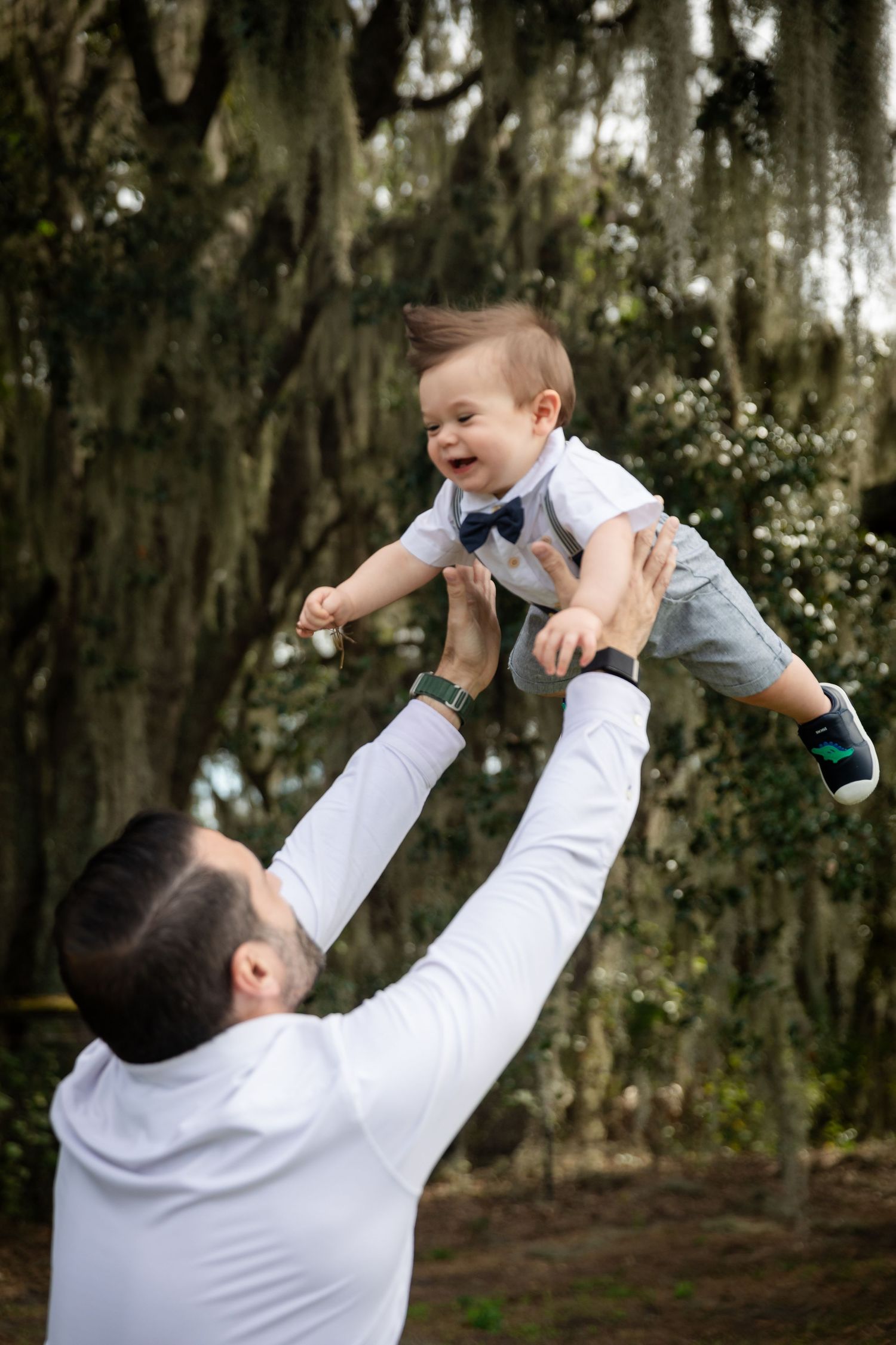 A joyful father lifts his laughing baby boy in the air outdoors beneath moss-draped oak trees.
