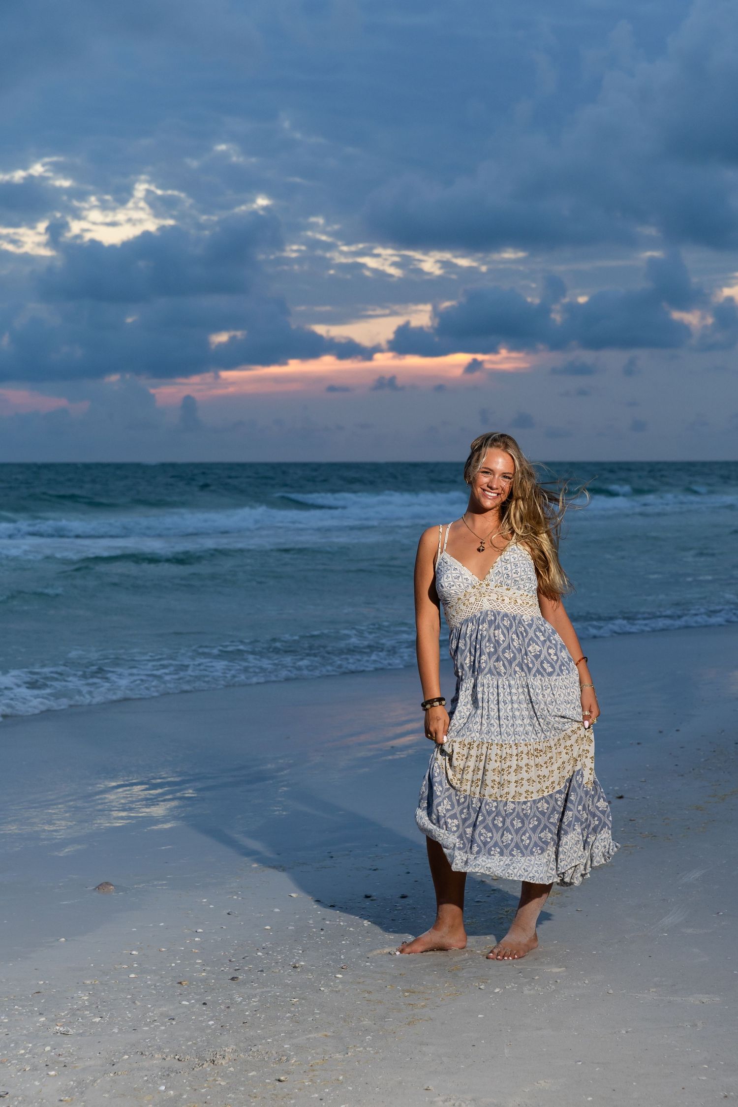 Smiling woman in boho blue dress walking barefoot on sandy beach at sunset with dramatic cloudy sky.