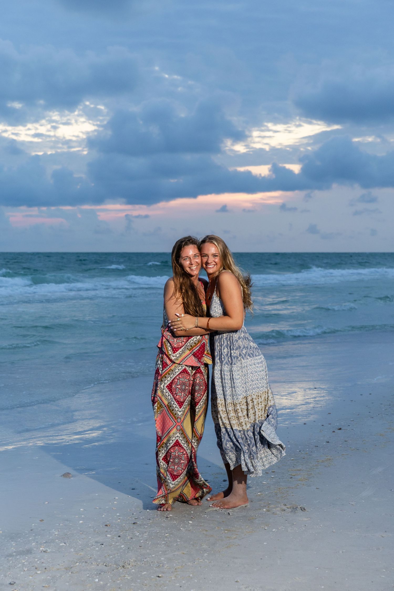 Two women in colorful maxi dresses embrace and smile on a sandy beach at sunset with ocean waves behind them.