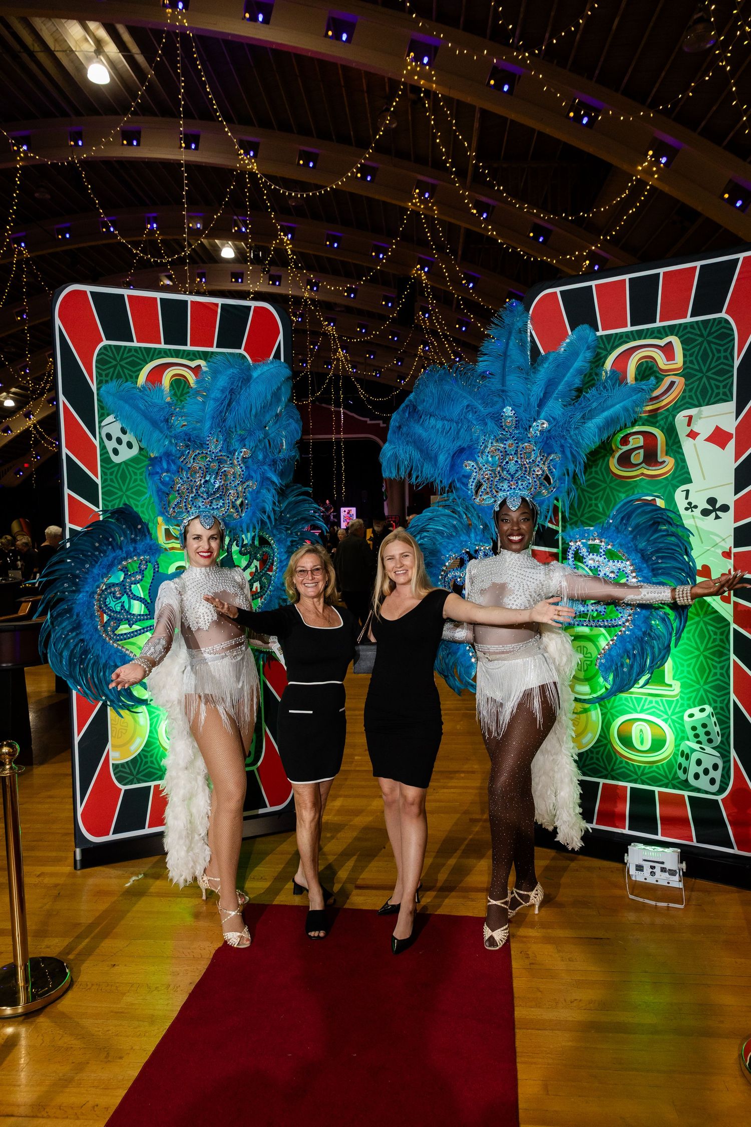 Two elegant showgirls in blue feathered headdresses pose with two women at a casino night event.