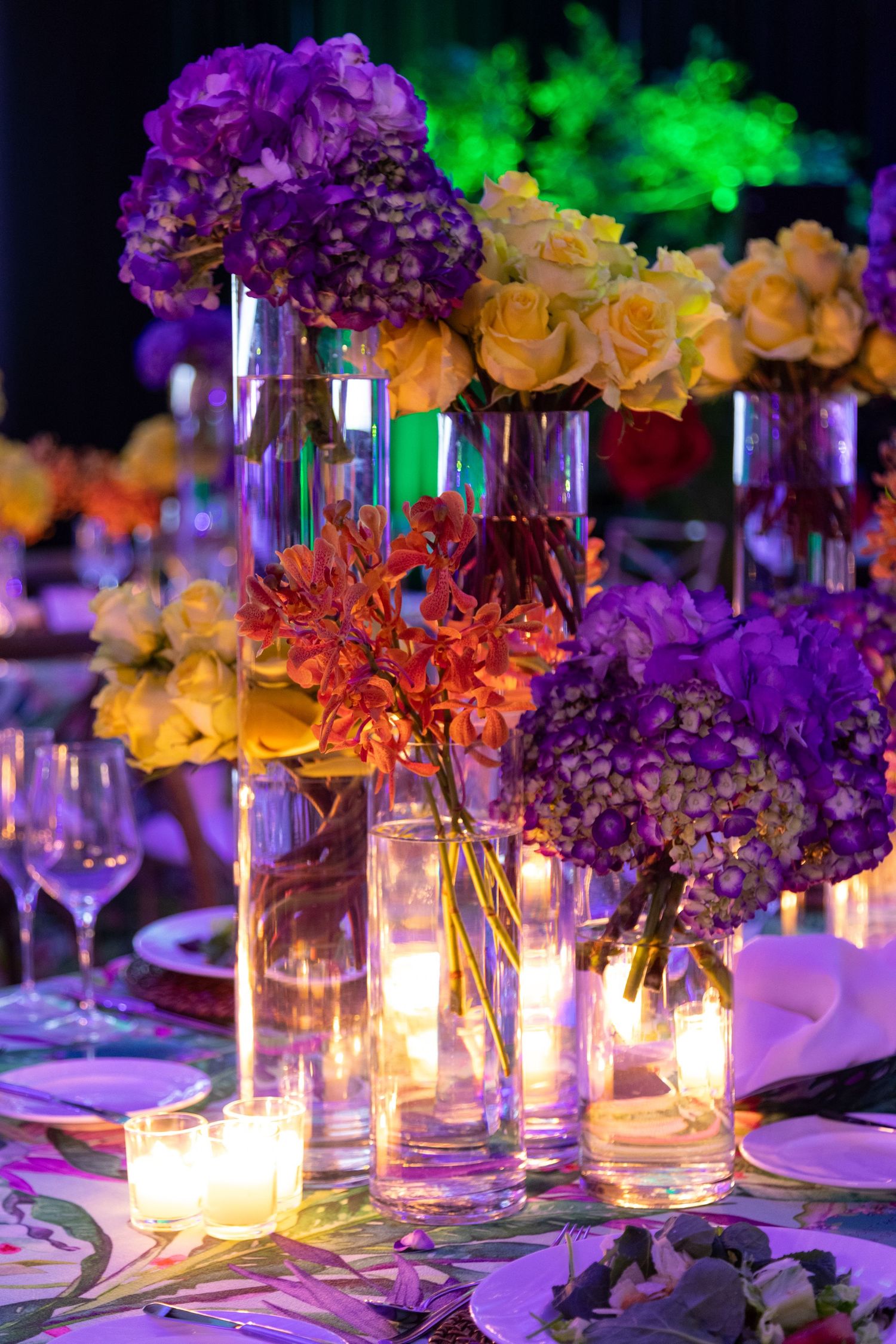 Elegant event table centerpiece with purple hydrangeas, yellow roses, and red flowers in glass vases with glowing candles.