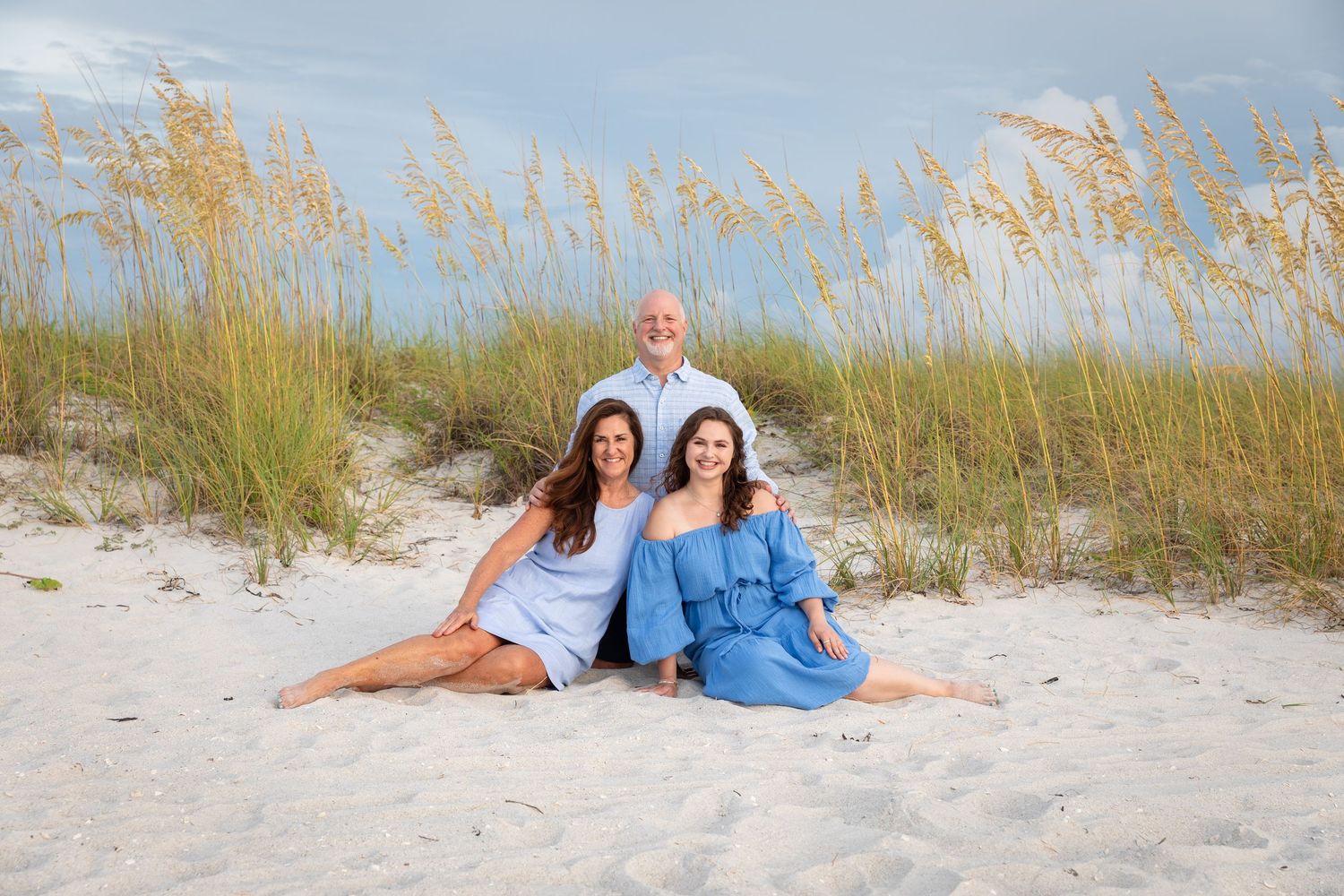 Family of three posing on white sand beach with sea oats, two women in blue dresses seated, man standing behind them.