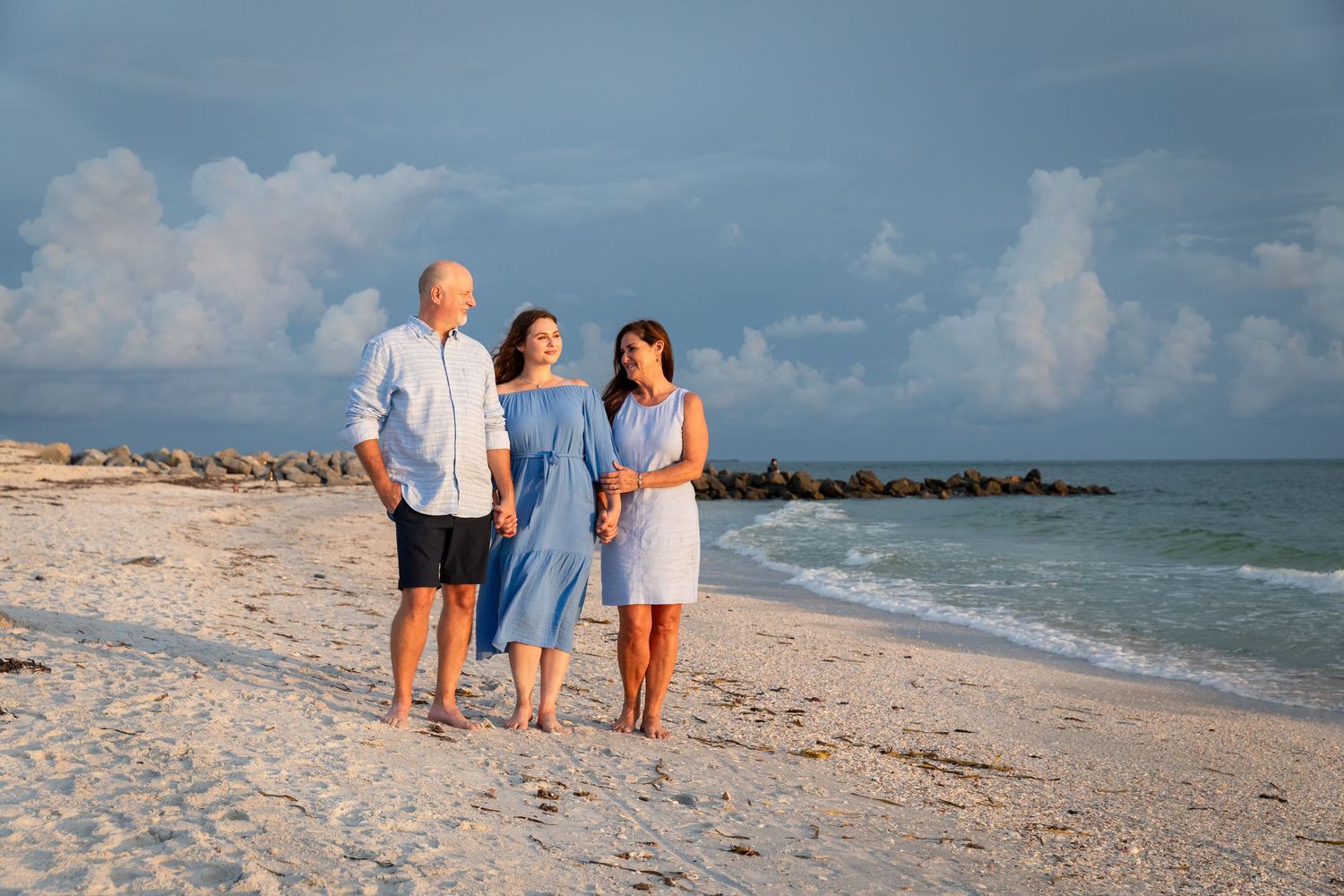 Family of three walking on a sandy beach at sunset, wearing blue outfits with ocean waves and cloudy sky.