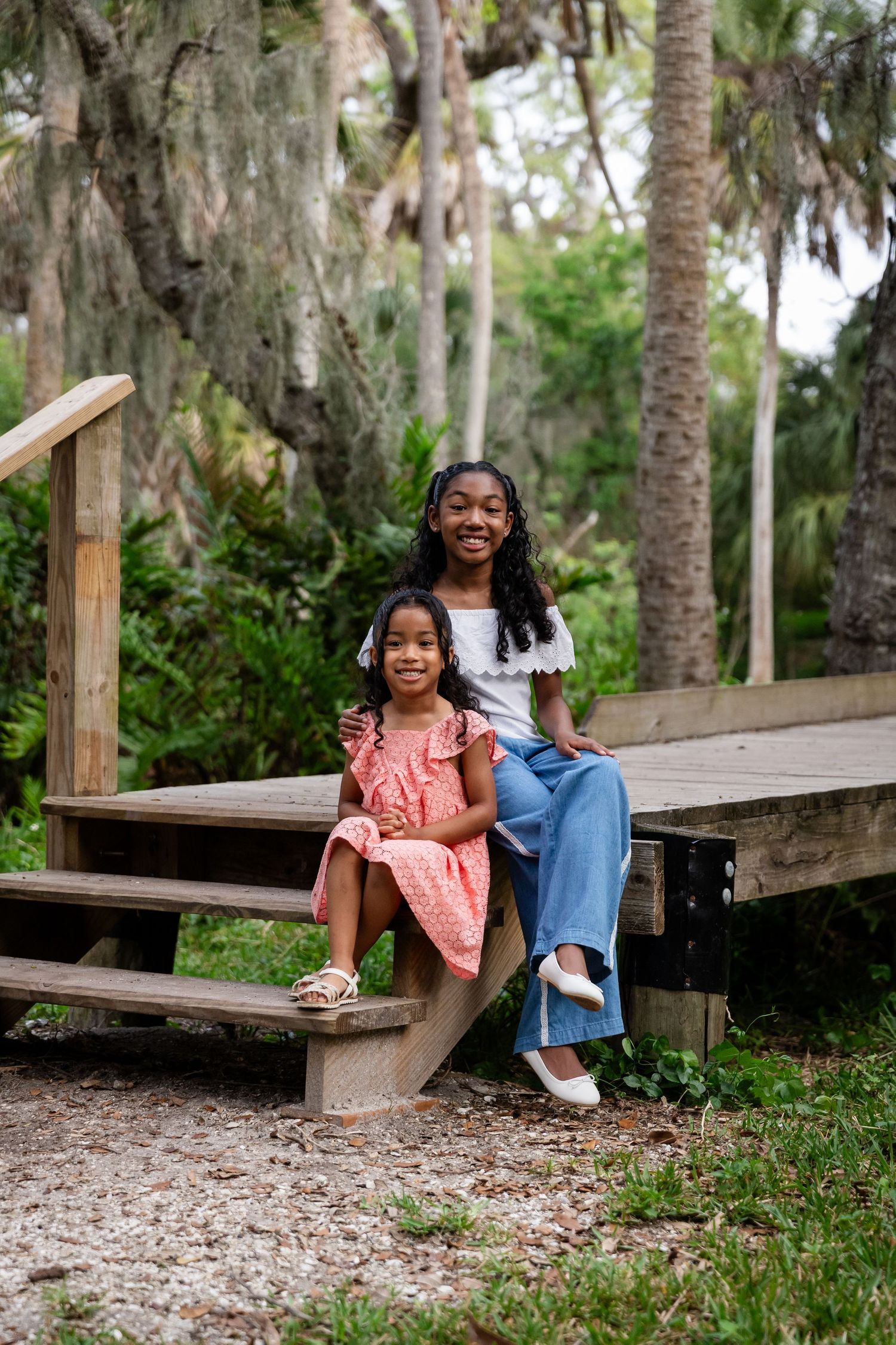 Two smiling girls sitting on wooden steps surrounded by lush greenery, one in a pink dress, one in jeans and white top.