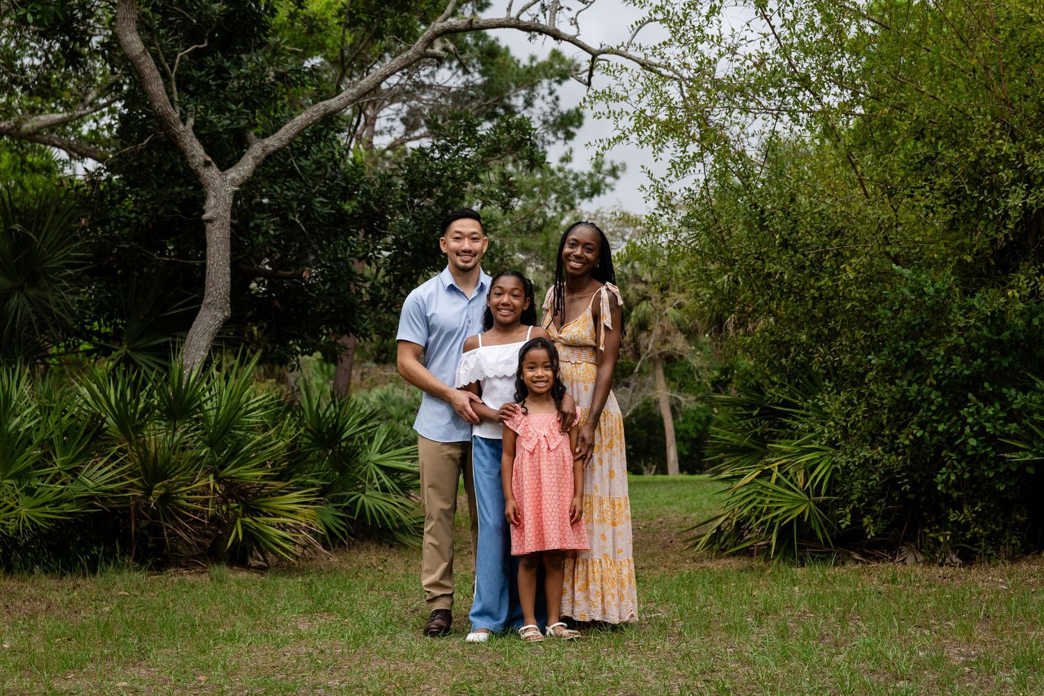 Family of four smiling together outdoors in a lush green park setting for a portrait photo.