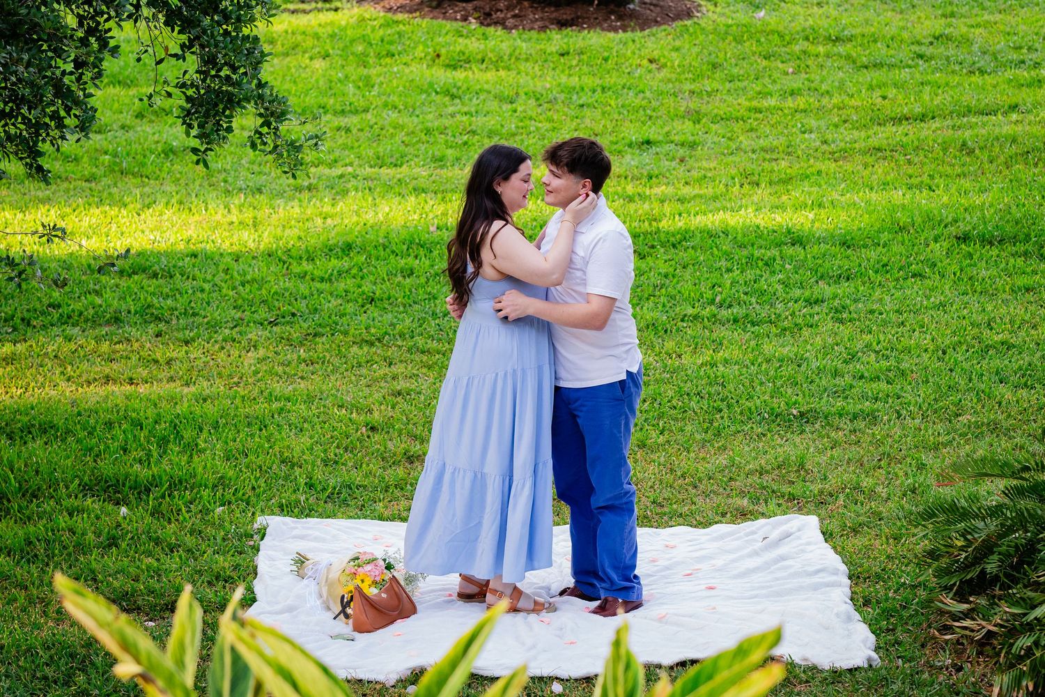 Couple sharing a romantic kiss on a white blanket in a lush green park, woman in blue dress, man in white shirt.