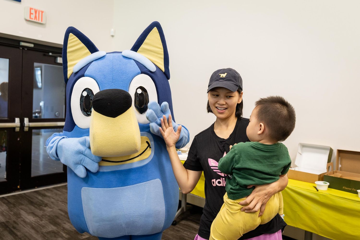 Woman holding toddler high-fiving a Bluey cartoon character mascot at a fun family event indoors.