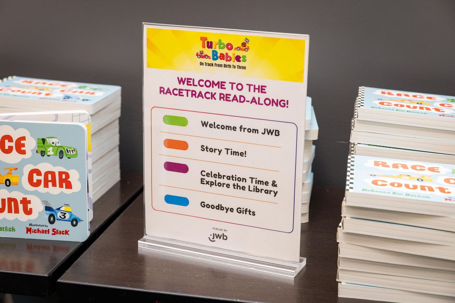 JWB Racetrack Read-Along welcome sign displaying event agenda including Story Time and Celebration at a children's library event.