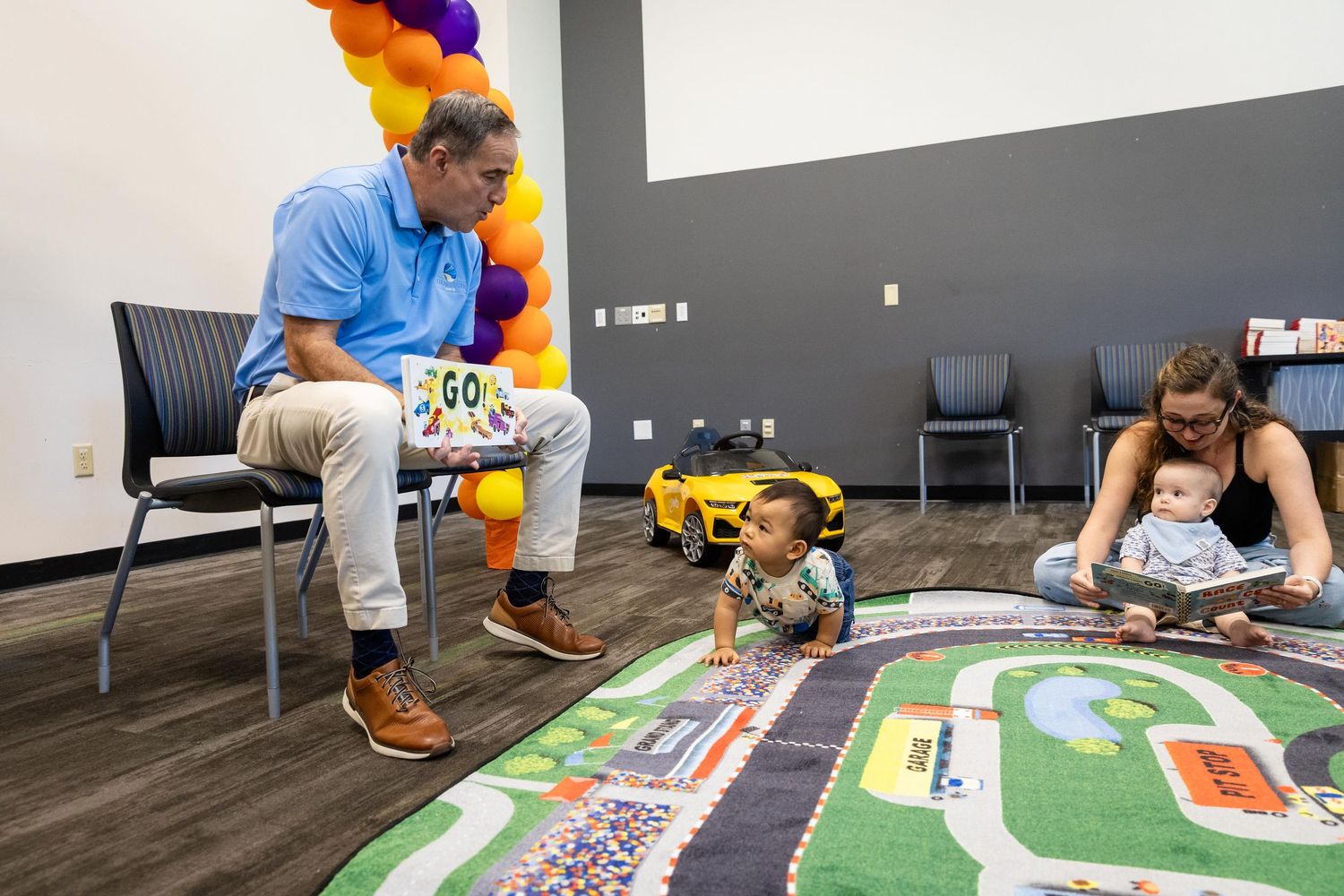 Man reads to toddler and mother on colorful road play mat in bright community room with balloon arch.