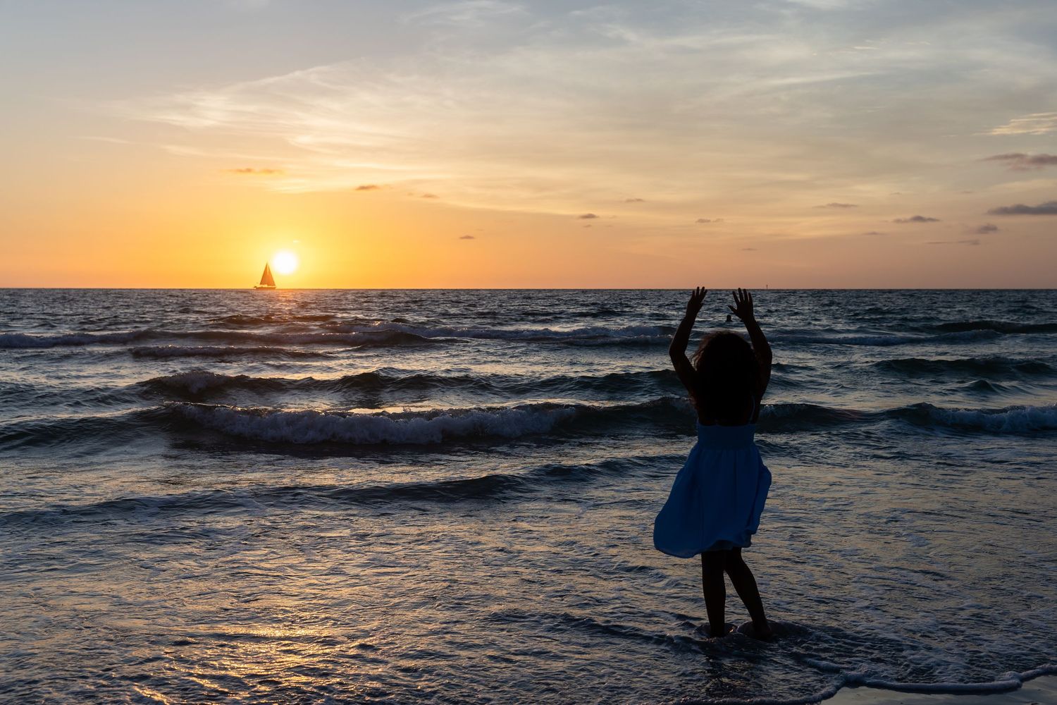 Silhouette of a child photographing a sailboat at sunset on a beautiful ocean beach with golden sky.