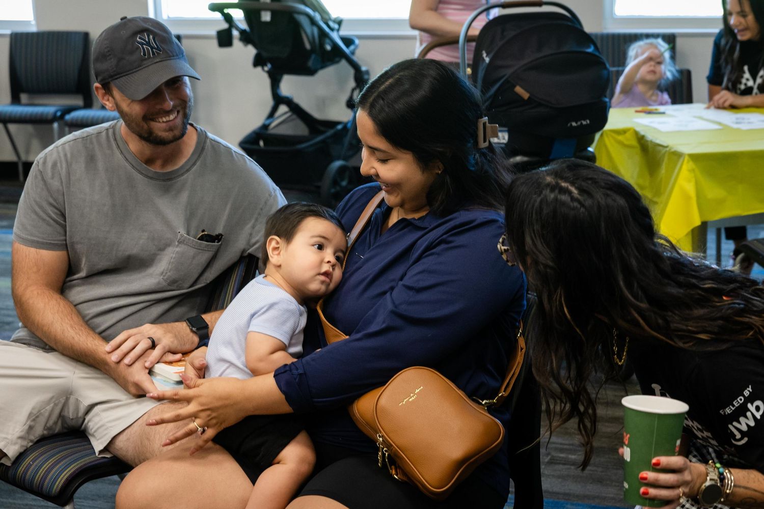 A family gathering with a man, woman holding a baby, and another person interacting warmly in a bright community room.