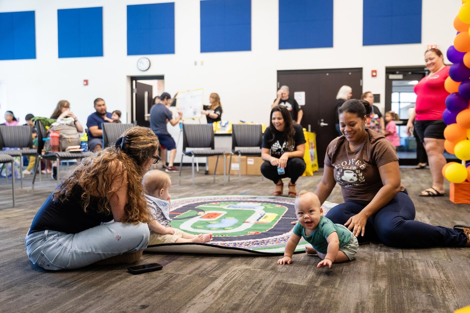 Two mothers and babies play on a colorful mat on a wooden floor during an indoor community playgroup event.