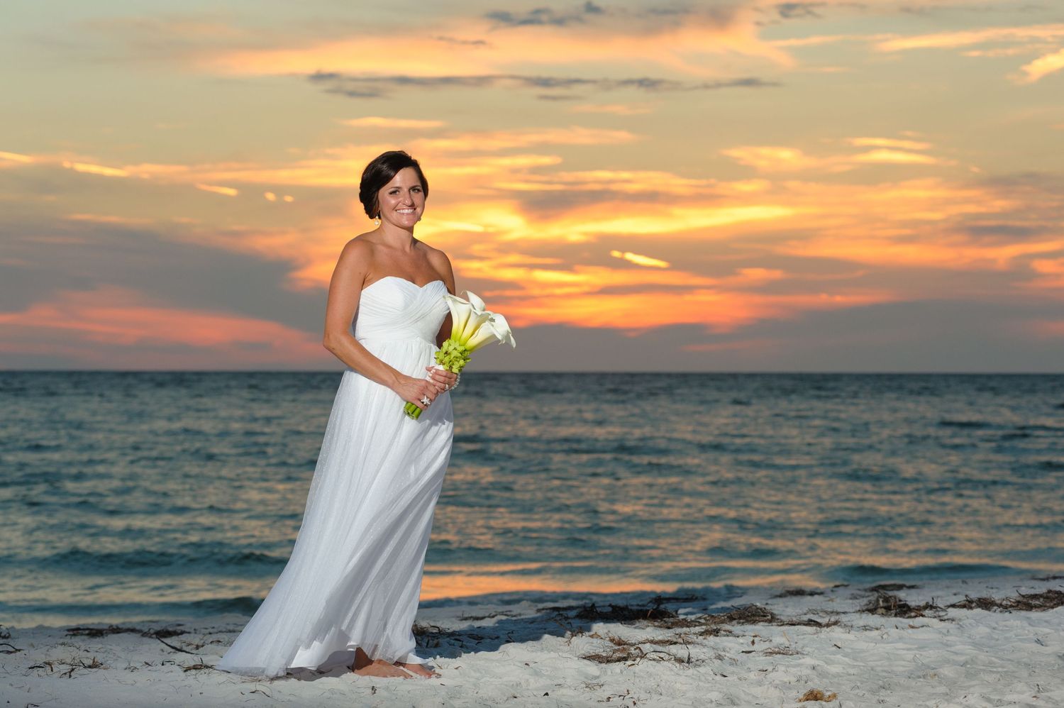 Elegant bride in a white gown holding calla lilies posing on the beach at sunset.