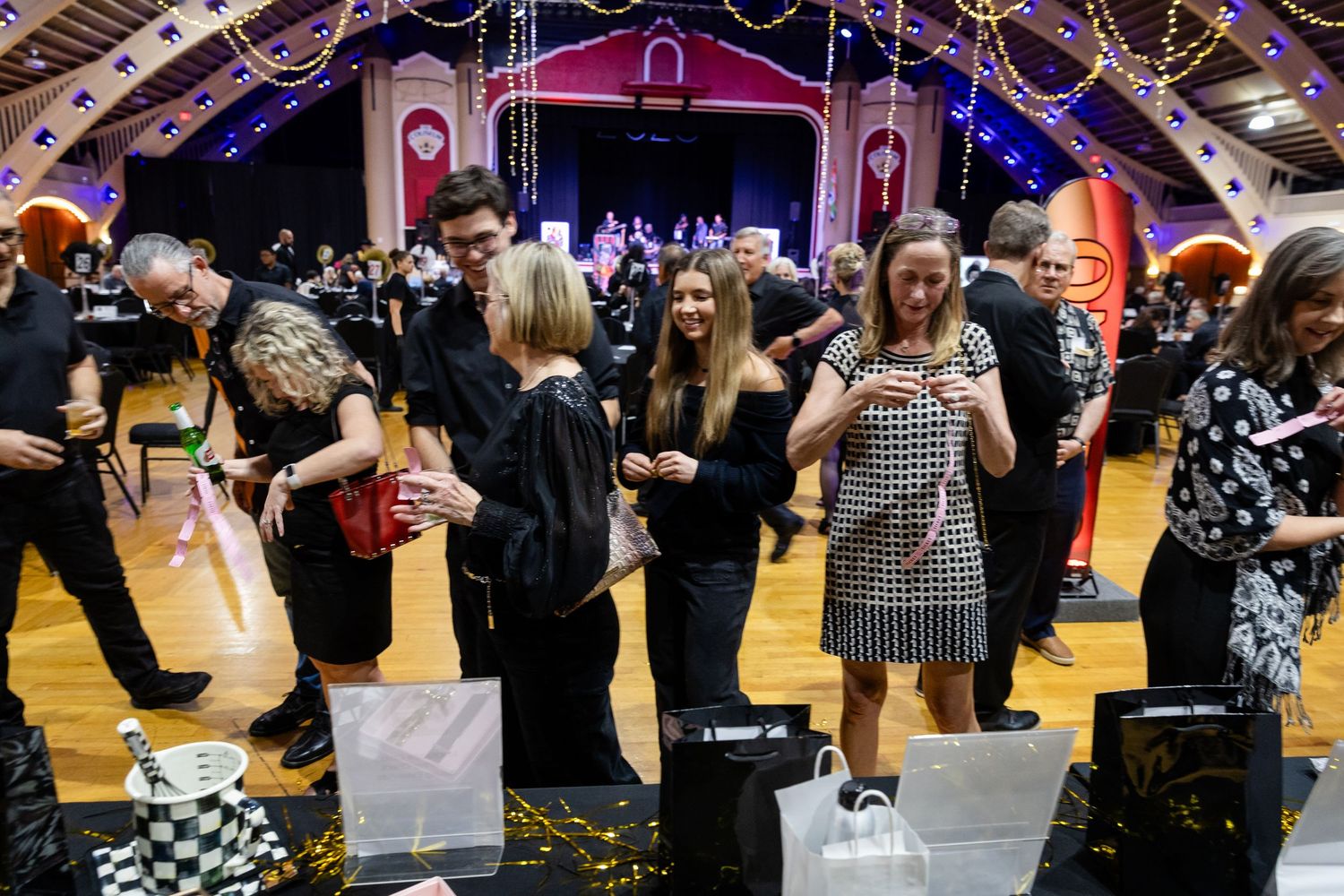 Guests dressed in black mingle around auction tables in a decorated event hall with a stage in background.