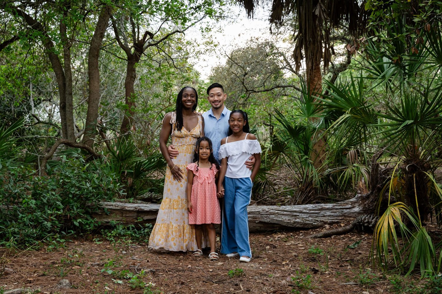 Happy interracial family of four posing together in a lush green wooded outdoor setting.