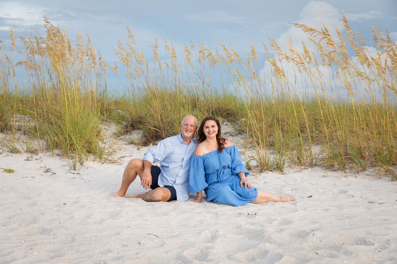 Couple sitting together on white sand beach dunes with sea oats, both wearing blue outfits, smiling.