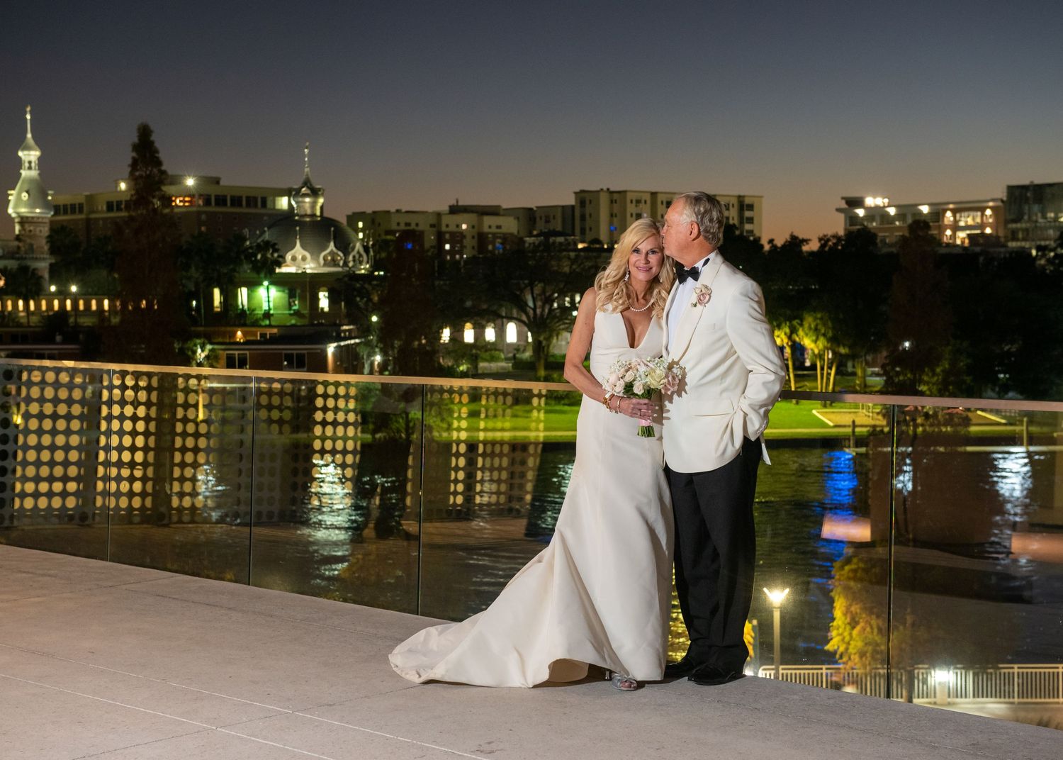 Couple sharing a romantic kiss on a rooftop terrace at night with a glittering city skyline backdrop at their wedding.