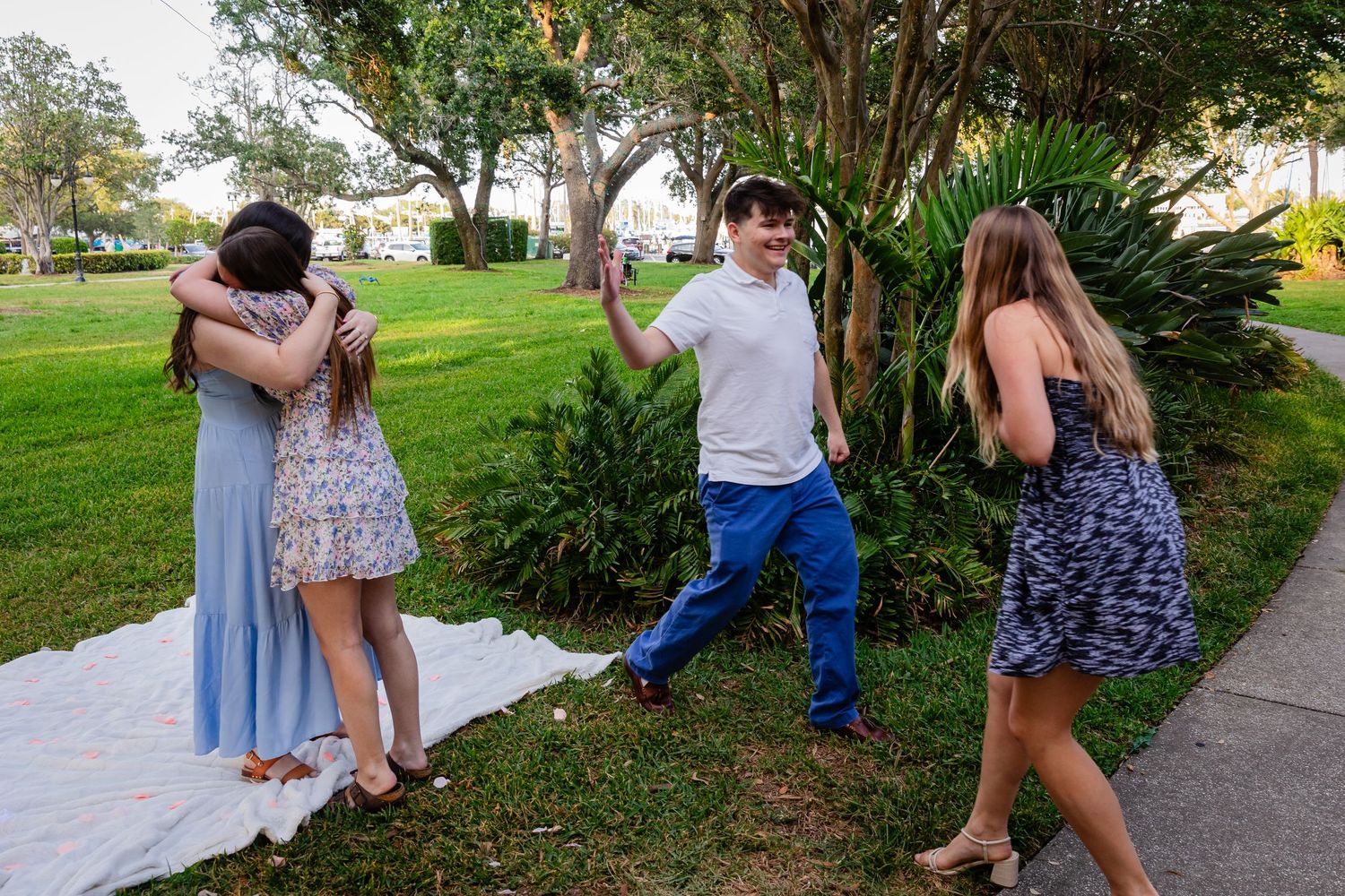 Four people hugging and greeting each other on a lawn beside a white blanket and lush green shrubs.