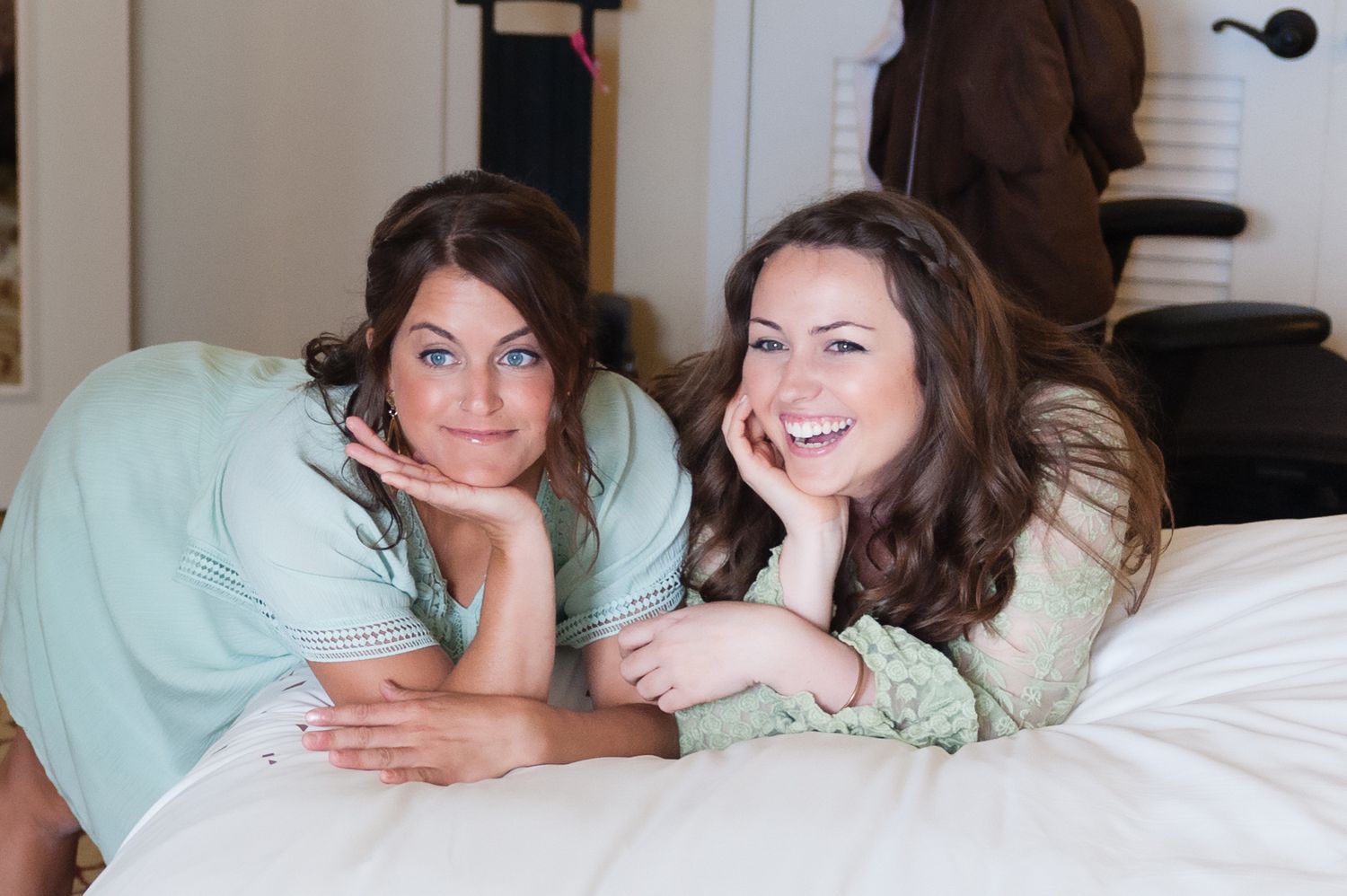 Two smiling women lying on a white bed, wearing light green tops, posing with hands on chins.