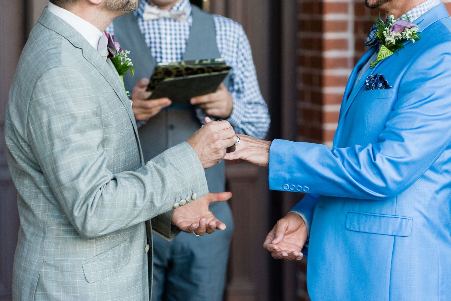 Two grooms exchange rings at a wedding ceremony, one in gray suit, one in blue suit, with officiant behind them.