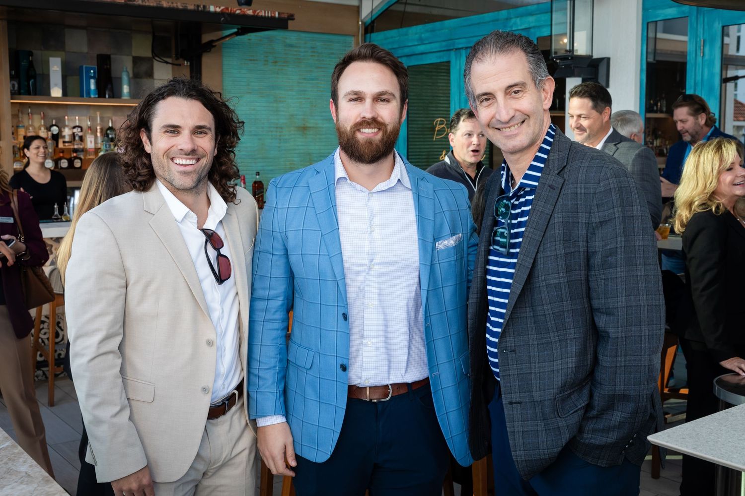 Three smiling men in blazers pose together at an indoor-outdoor social event with guests in the background.