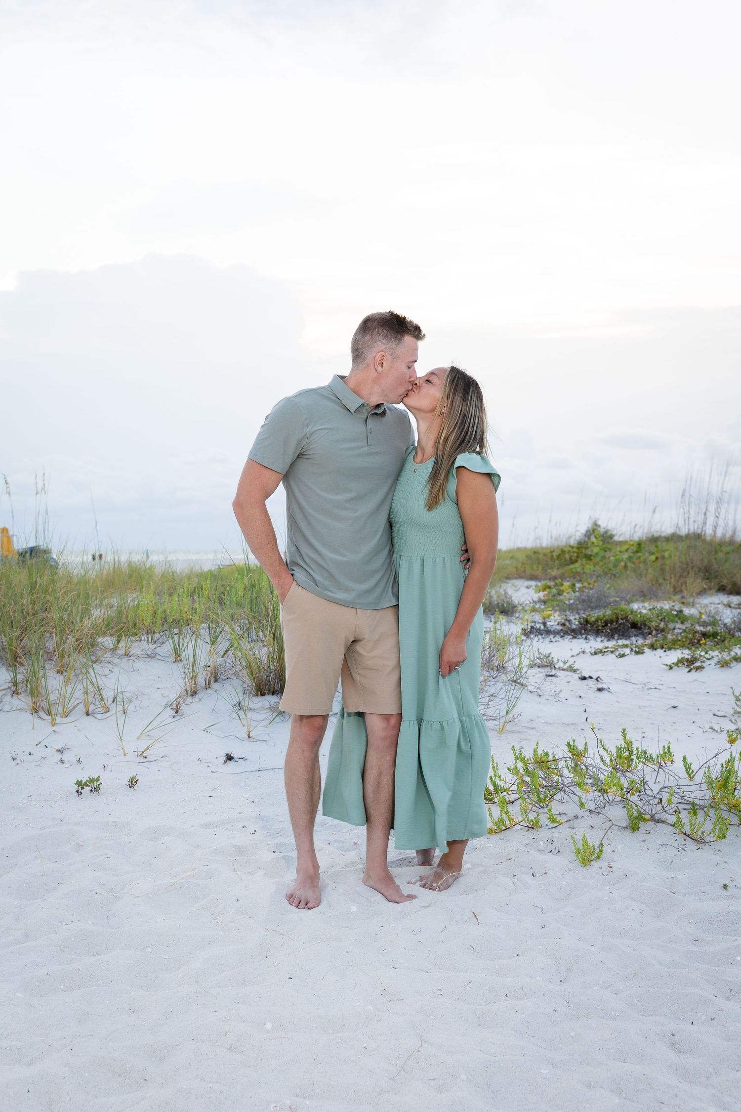 Couple kissing on white sand beach, man in gray polo and khaki shorts, woman in teal maxi dress, ocean backdrop.