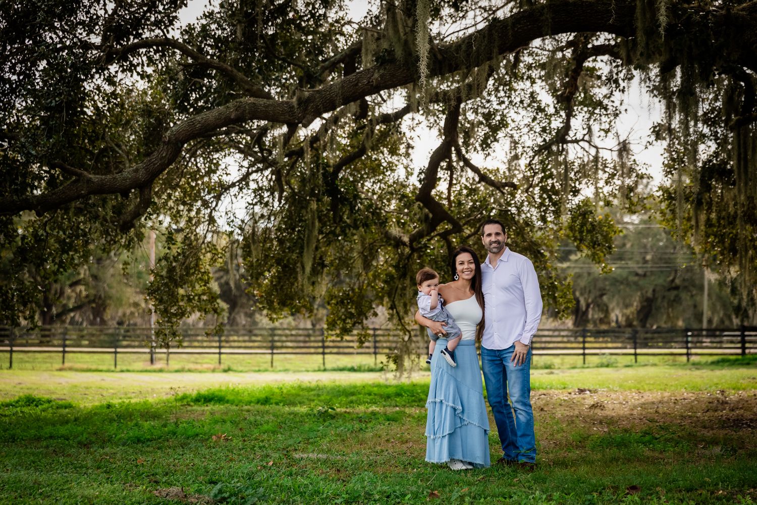 A young family with an infant stands together on a green lawn beneath a large oak tree on a farm.