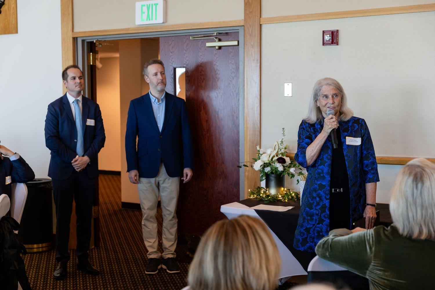 Woman speaking at podium with microphone at formal indoor event, two men standing nearby in professional attire.