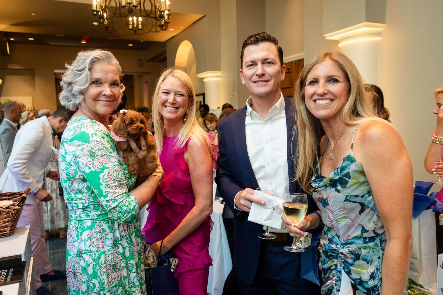 Four guests smiling and socializing at an elegant indoor event, one woman holding a small dog.
