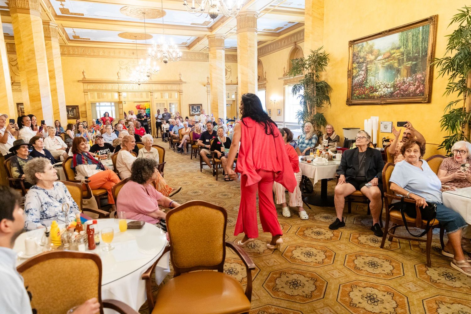 Performer in red dress walks through crowded elegant ballroom during a senior community event.