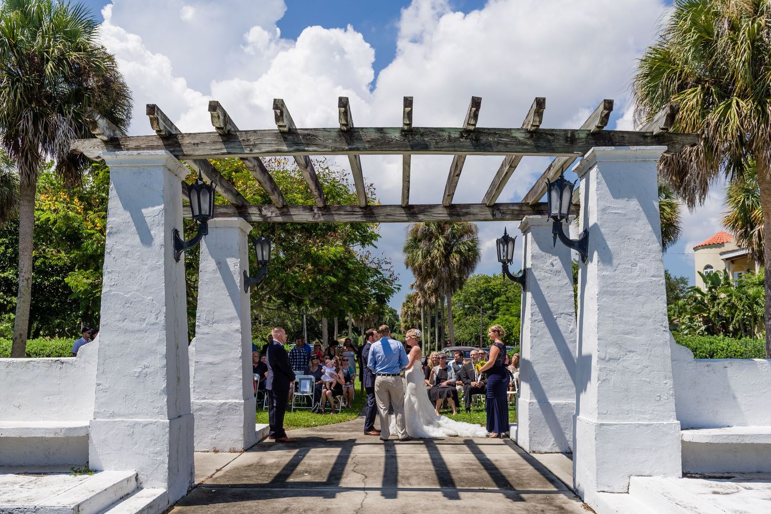 Outdoor wedding ceremony under a white pergola surrounded by palm trees on a sunny day.