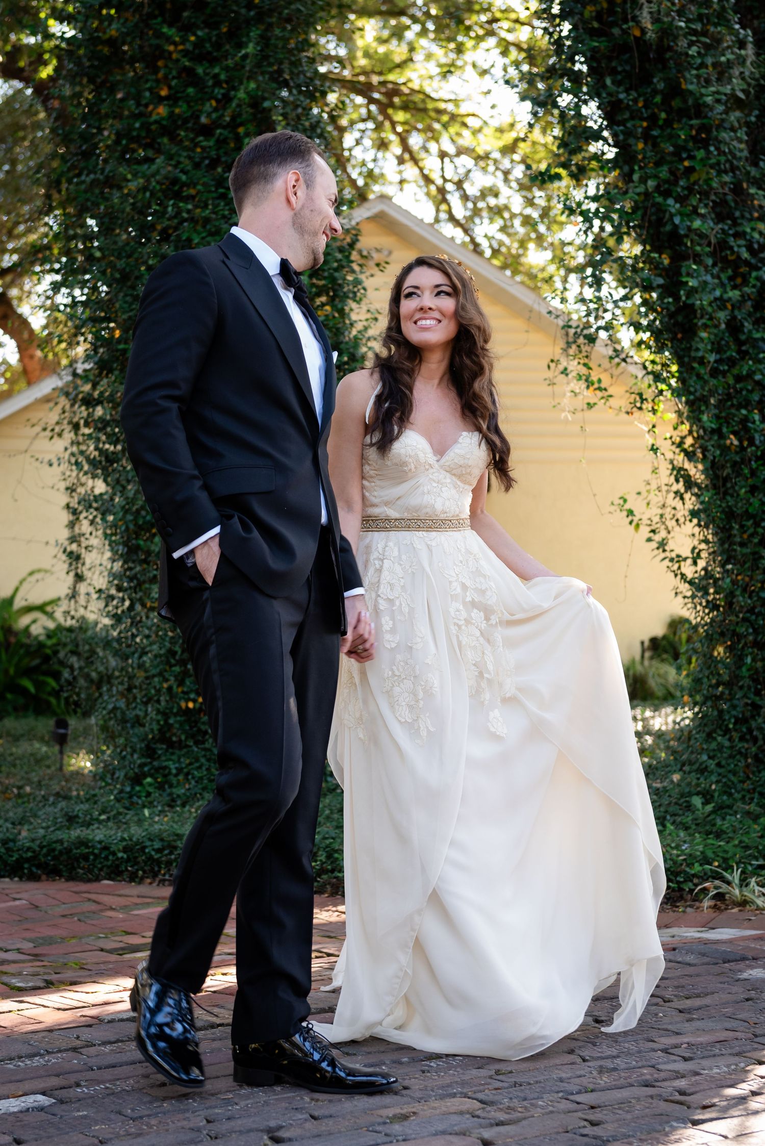 Bride in elegant white gown smiles at groom in black tuxedo on brick path surrounded by lush greenery.