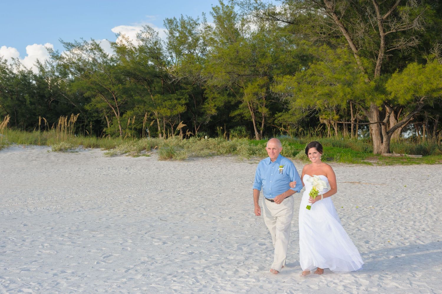 Bride and groom walking hand in hand on a white sandy beach with lush green trees in the background.