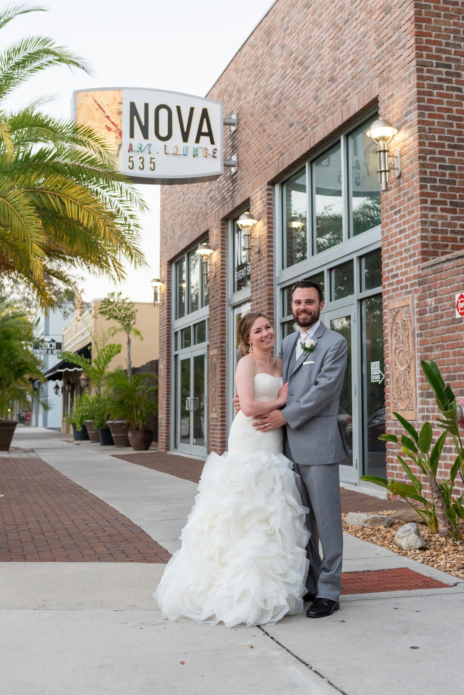 Smiling newlyweds pose together outside Nova Art Lounge 535 venue on their wedding day at dusk.