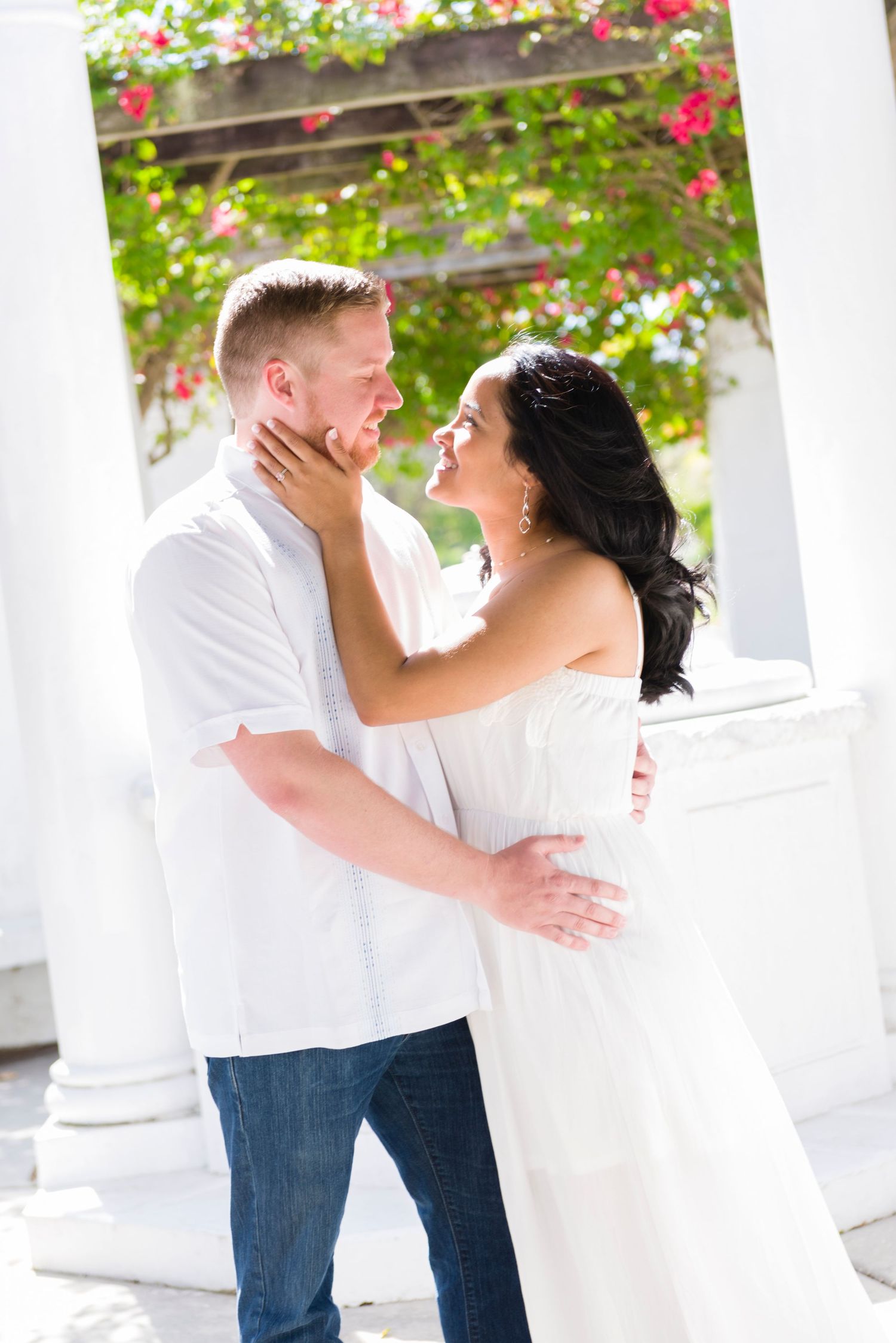 Romantic couple embracing under white columns with pink flowers, woman in white dress touching man's face outdoors.