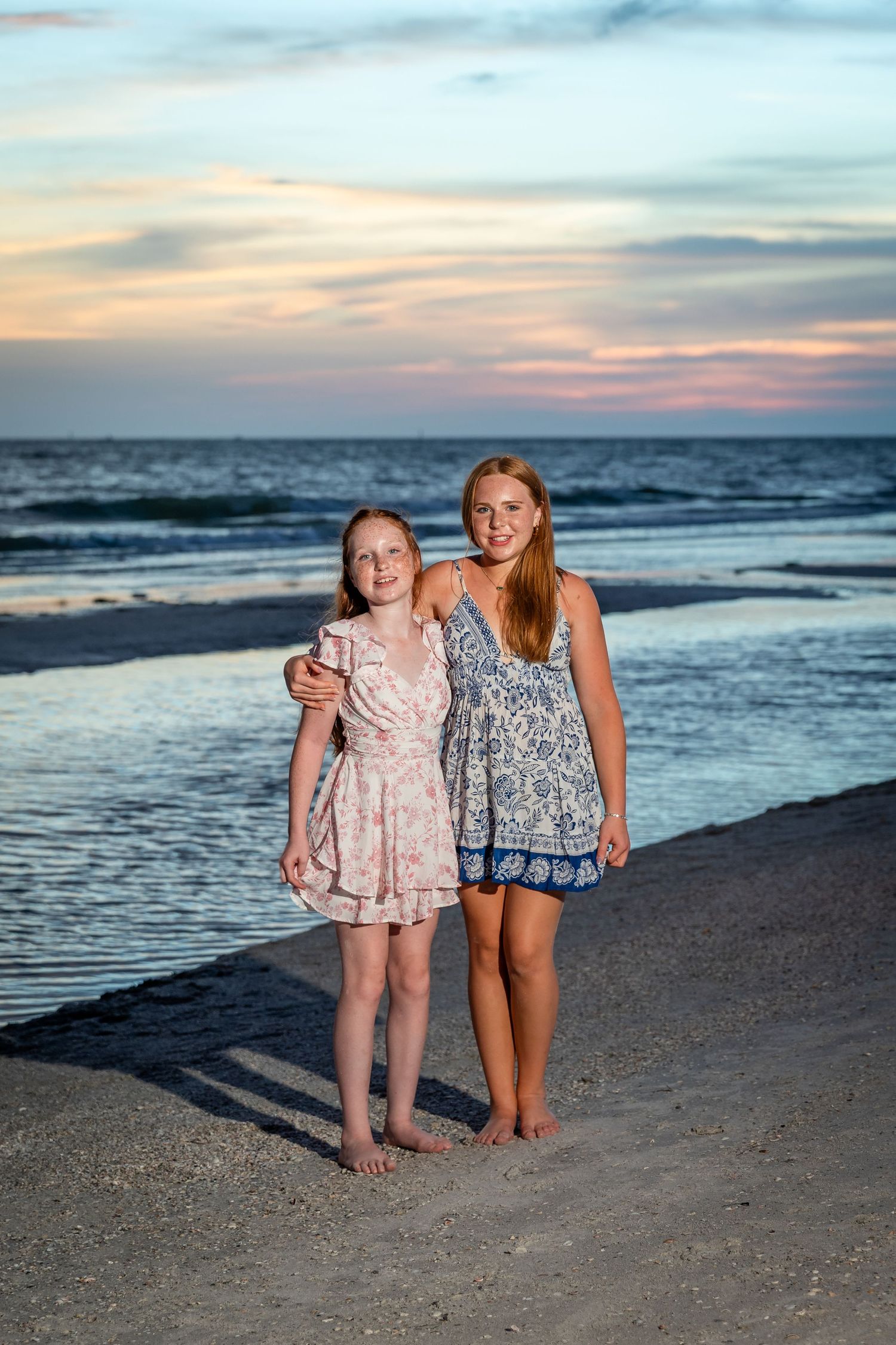 Two girls in summer dresses pose together on a sandy beach at sunset with ocean waves in the background.