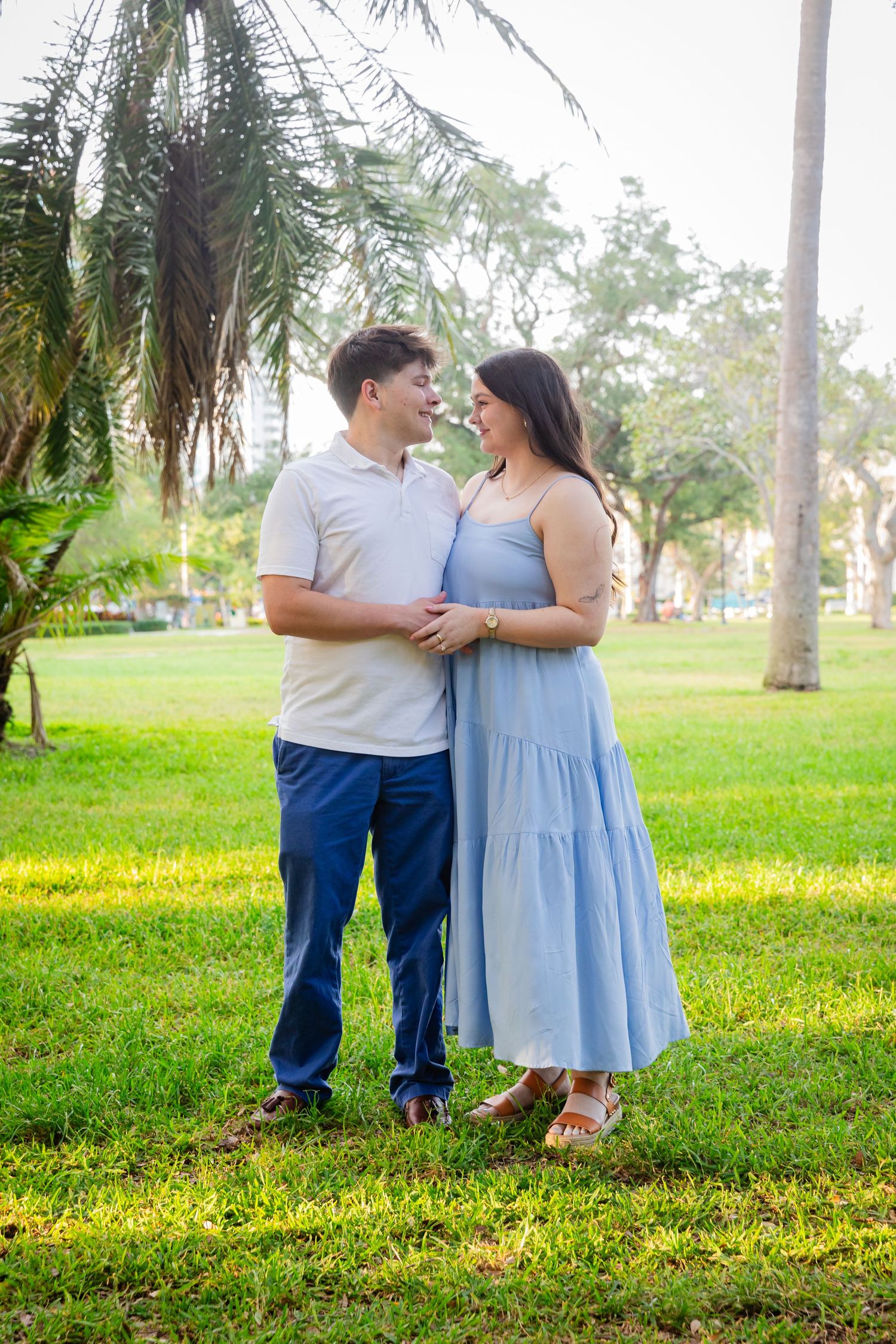 A couple in coordinating blue outfits smiling at each other while standing on green grass near palm trees.