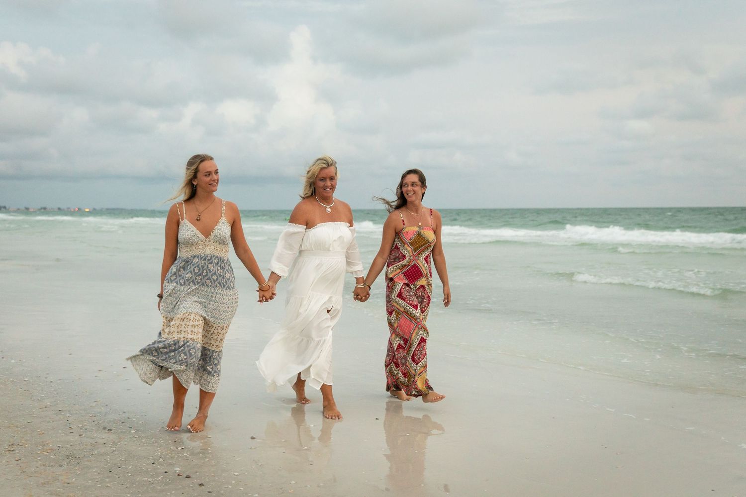 Three women in colorful dresses walk hand-in-hand along a sandy beach with ocean waves in the background.