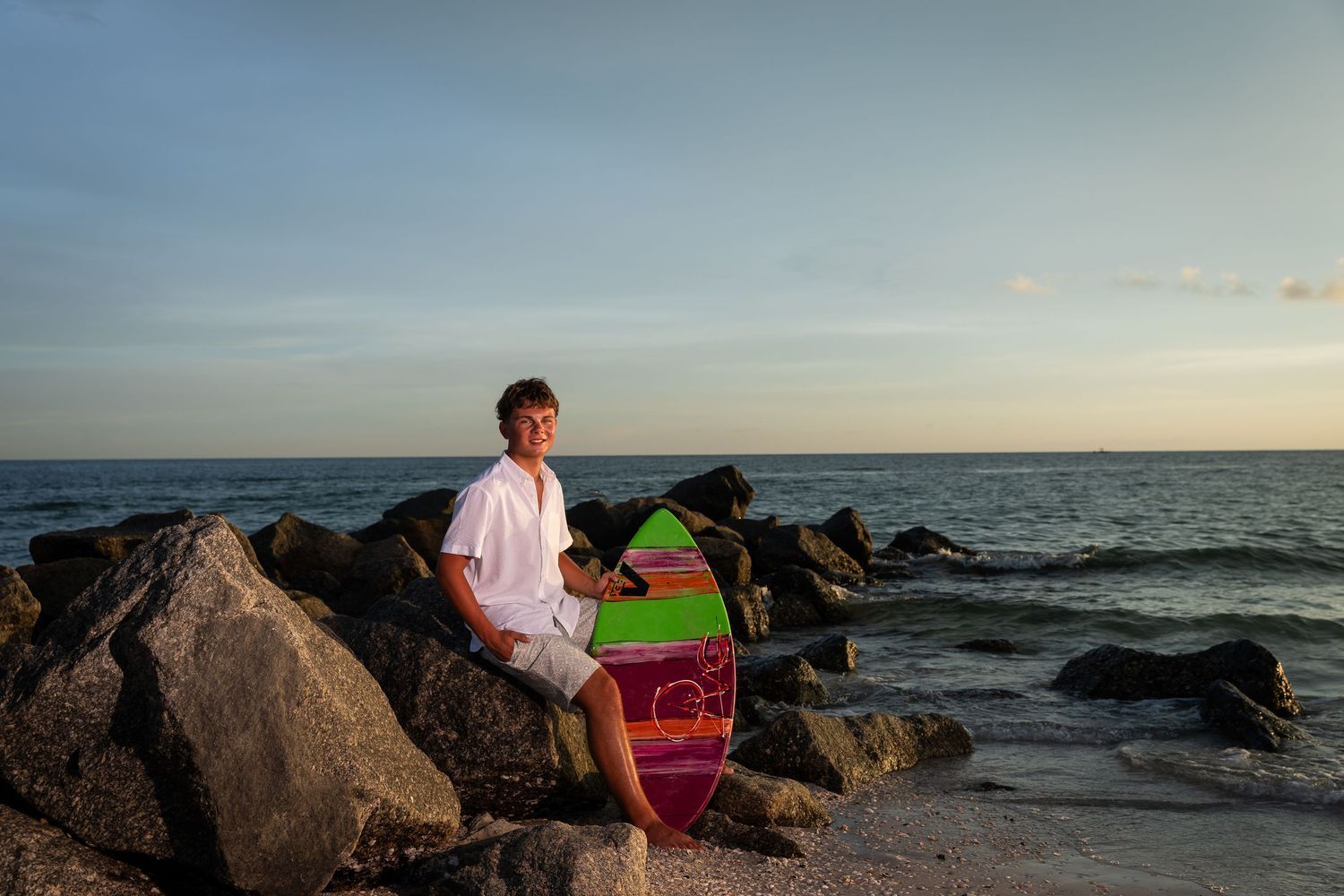 Teen boy sitting on rocks at sunset beach holding a colorful surfboard with ocean waves in background.