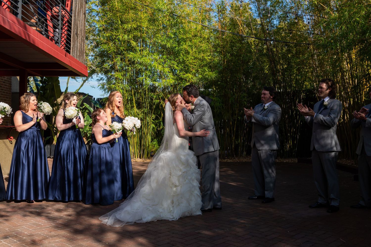 Bride and groom share first kiss surrounded by wedding party in bamboo garden.