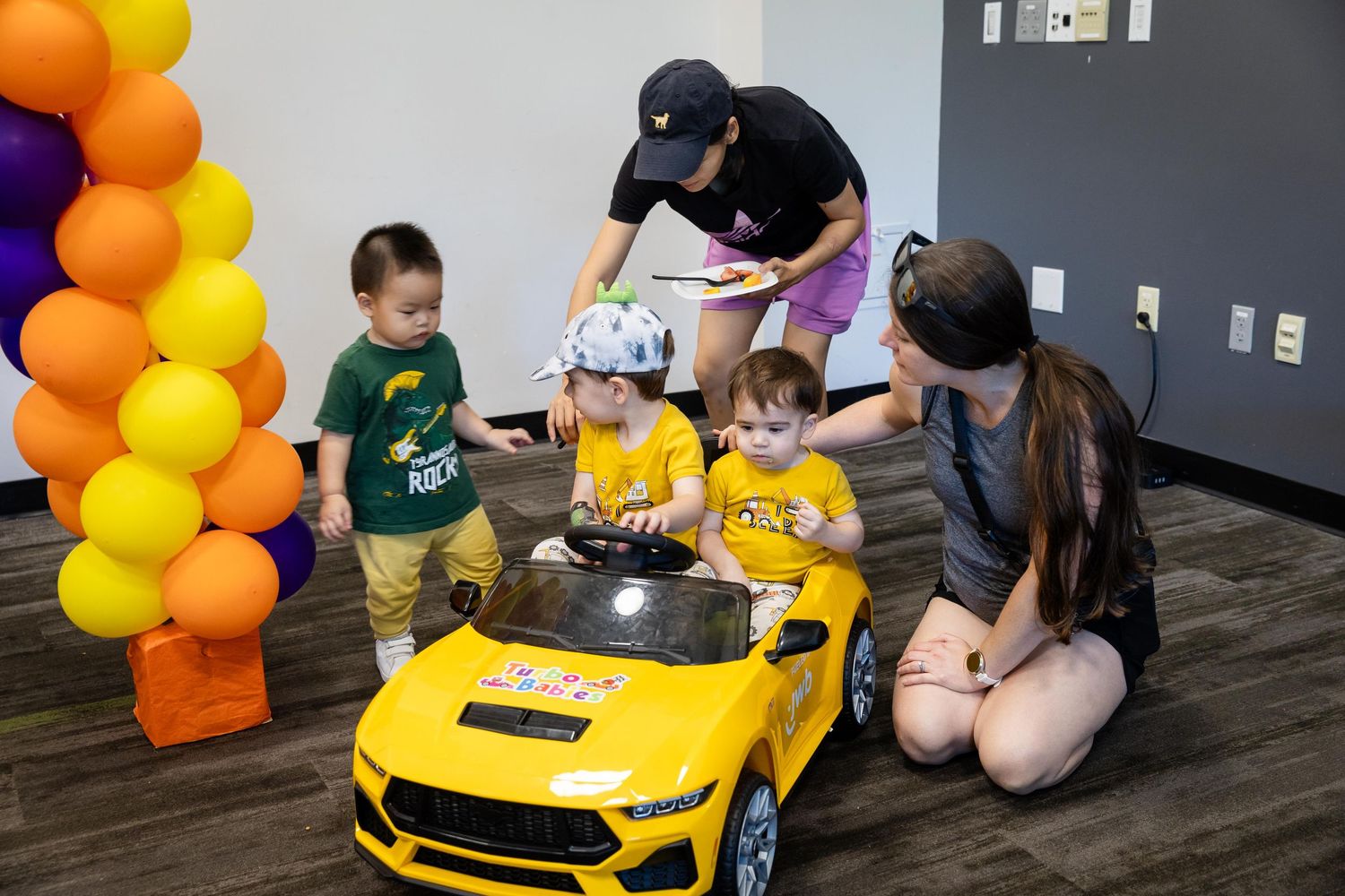Two toddlers in yellow shirts sit in a toy yellow Mustang car at a colorful balloon-decorated birthday party.