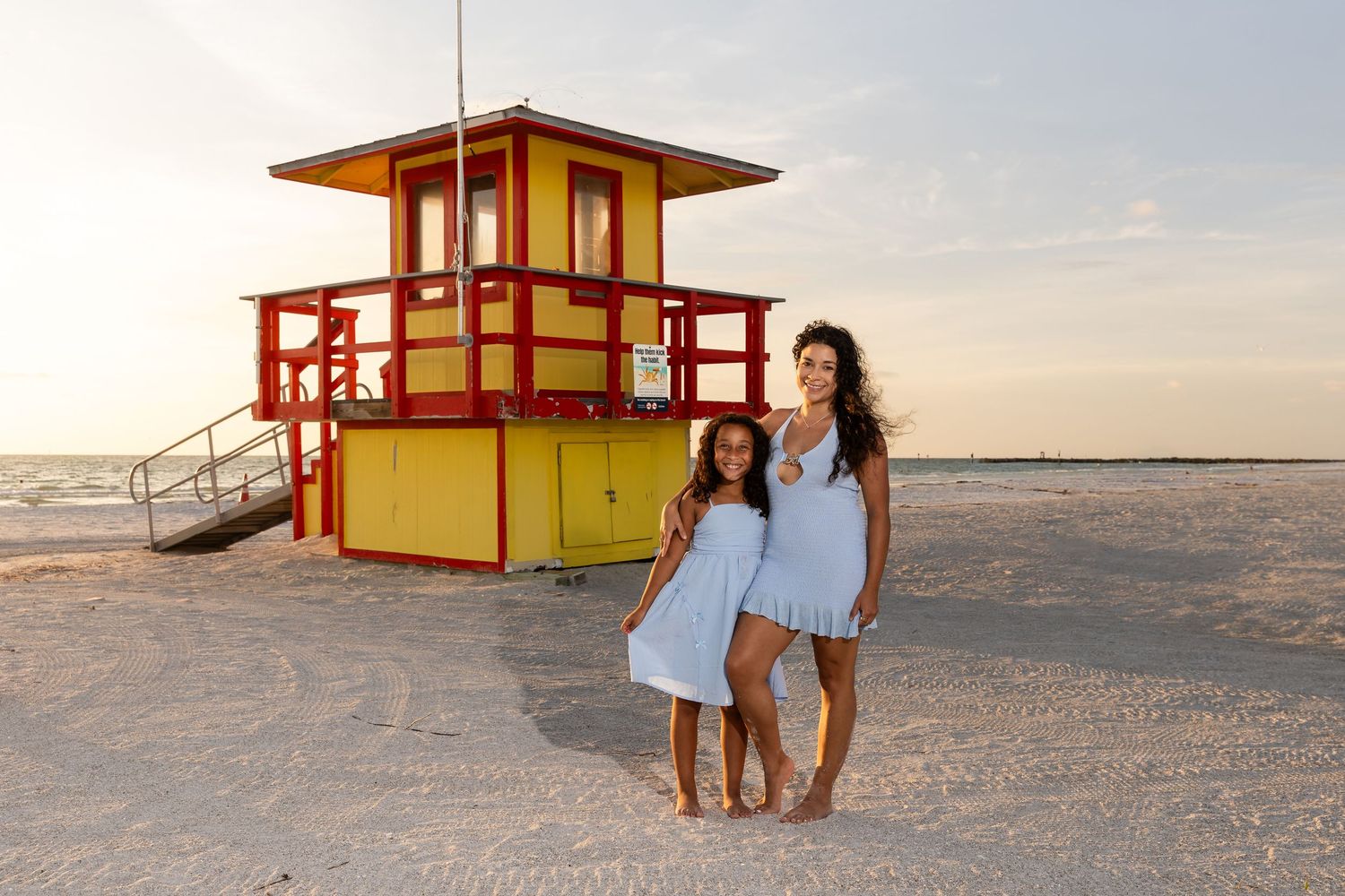 Woman and girl in blue dresses pose in front of colorful red and yellow lifeguard tower on sandy beach.