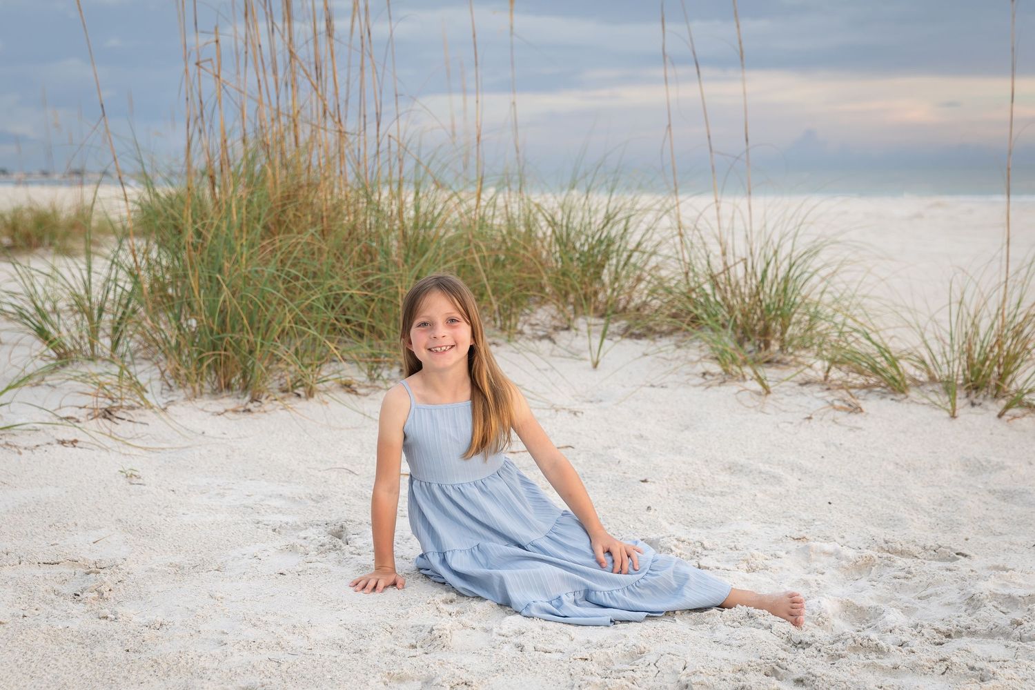 A smiling girl in a blue dress sits on white sand beach dunes with sea oats in the background.