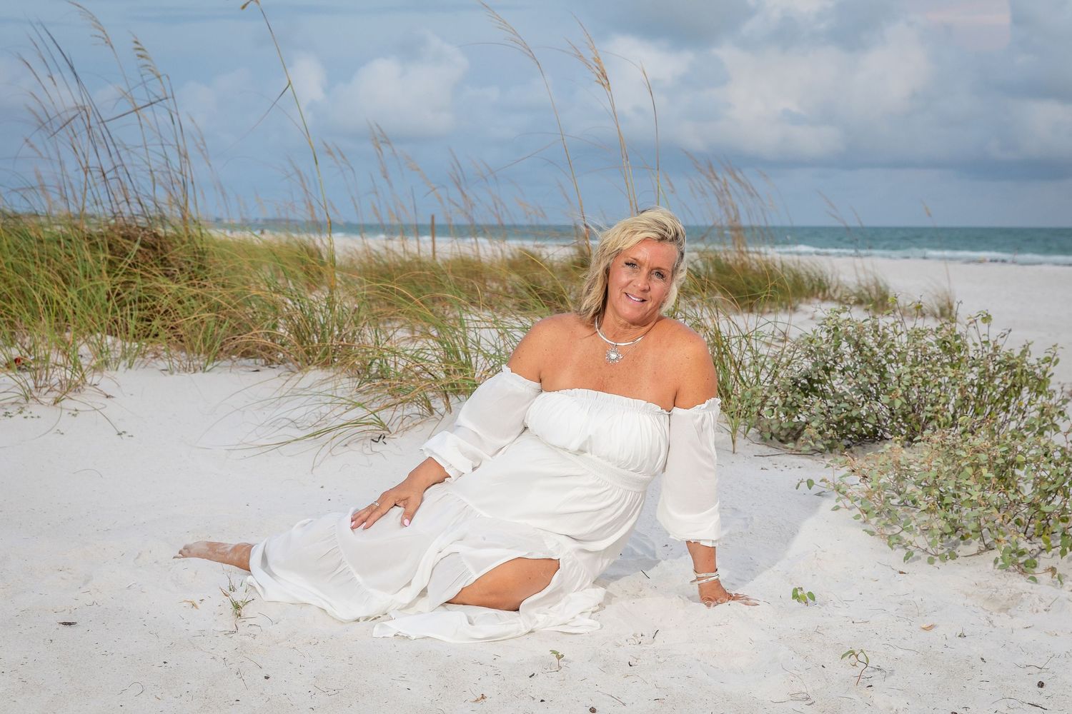 Blonde woman in white off-shoulder dress sitting gracefully on white sand dunes with sea oats.