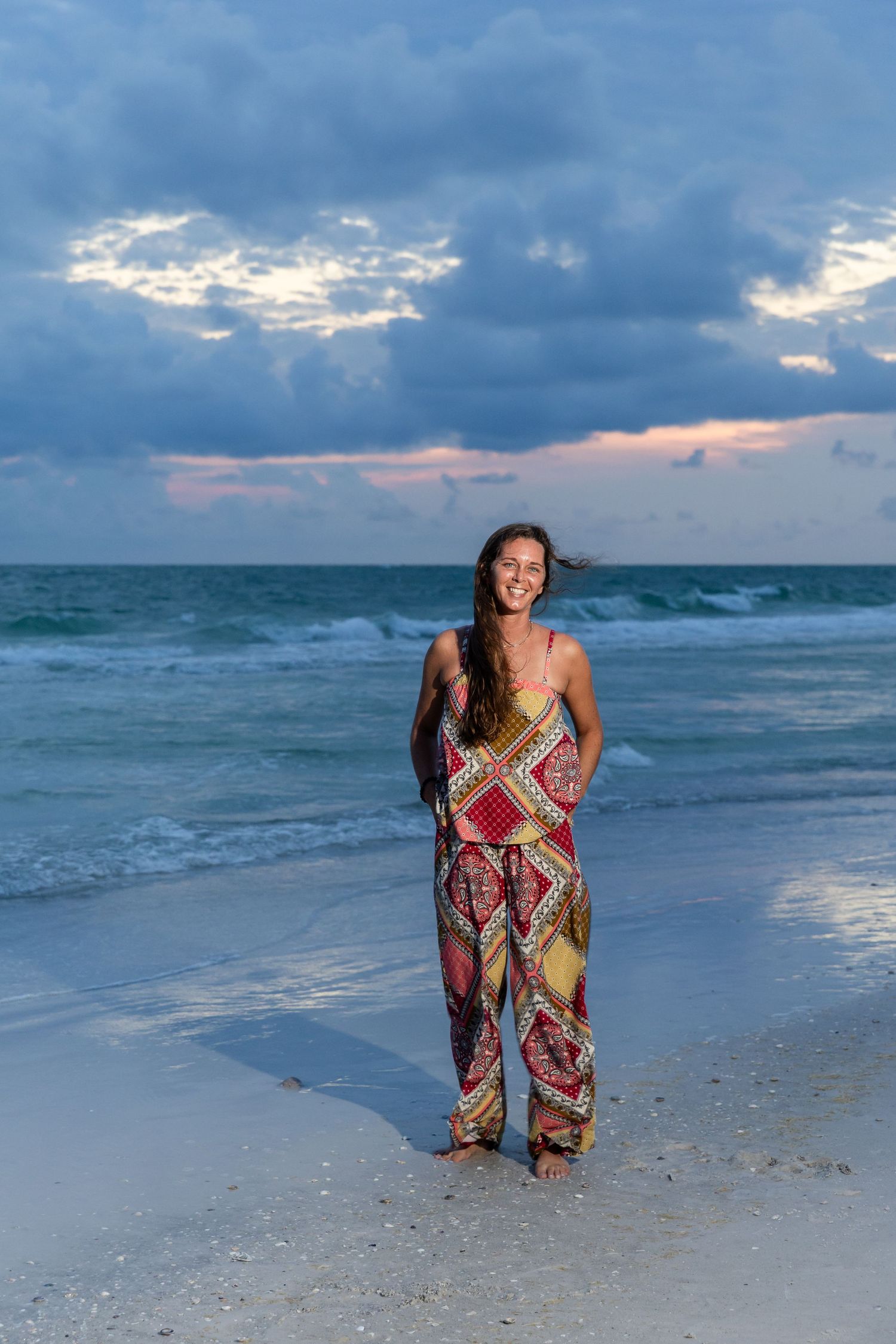 Woman in colorful patterned jumpsuit smiling on a white sand beach at sunset with dramatic cloudy sky.