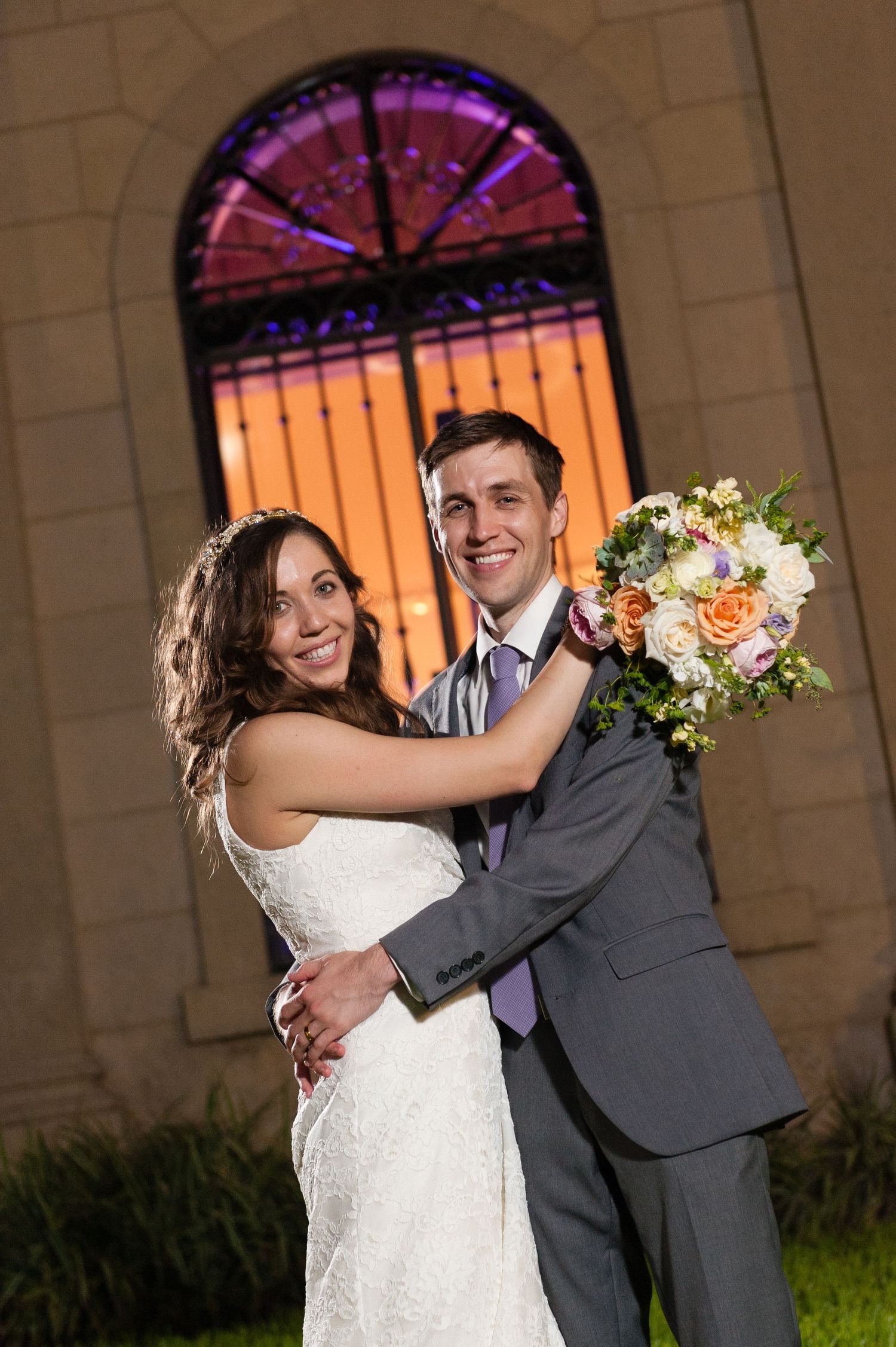 Smiling bride and groom pose together at night holding a bouquet in front of a stained glass window.