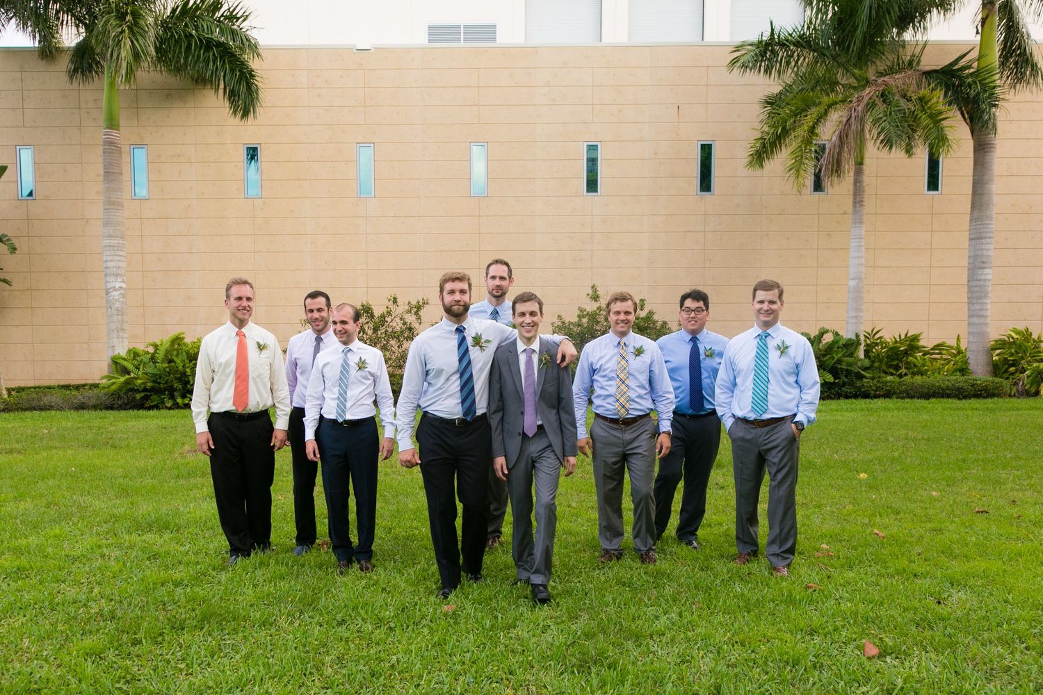Group of groomsmen in suits and colorful ties posing on green lawn with palm trees in background.